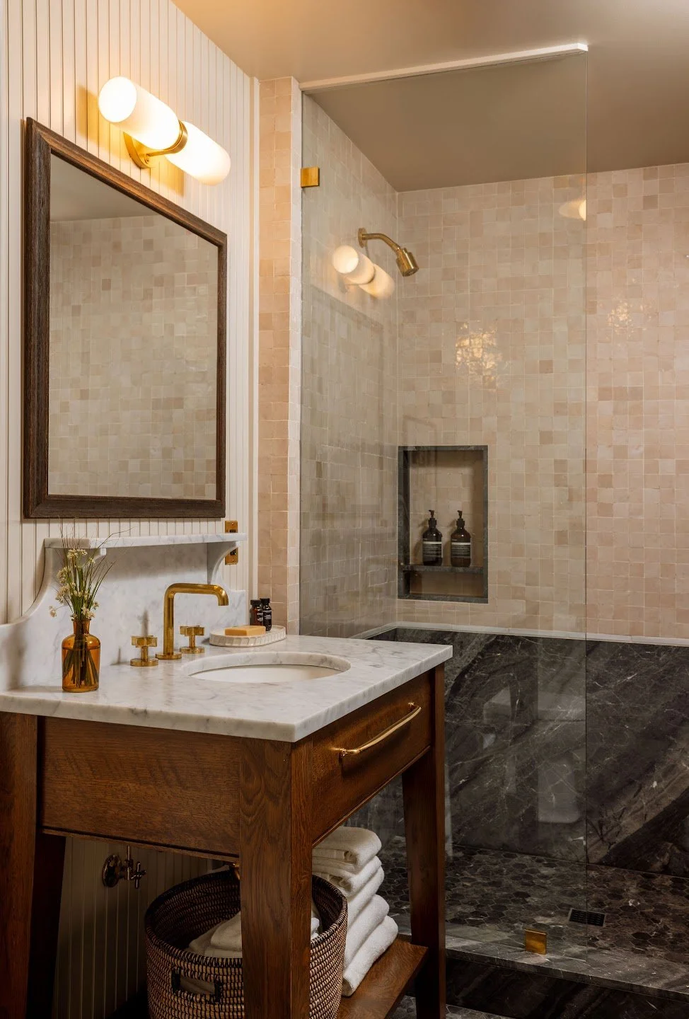 A bathroom with a vanity mirror, a marble countertop with a gold faucet, and a walk-in shower with beige tiles and a glass door.