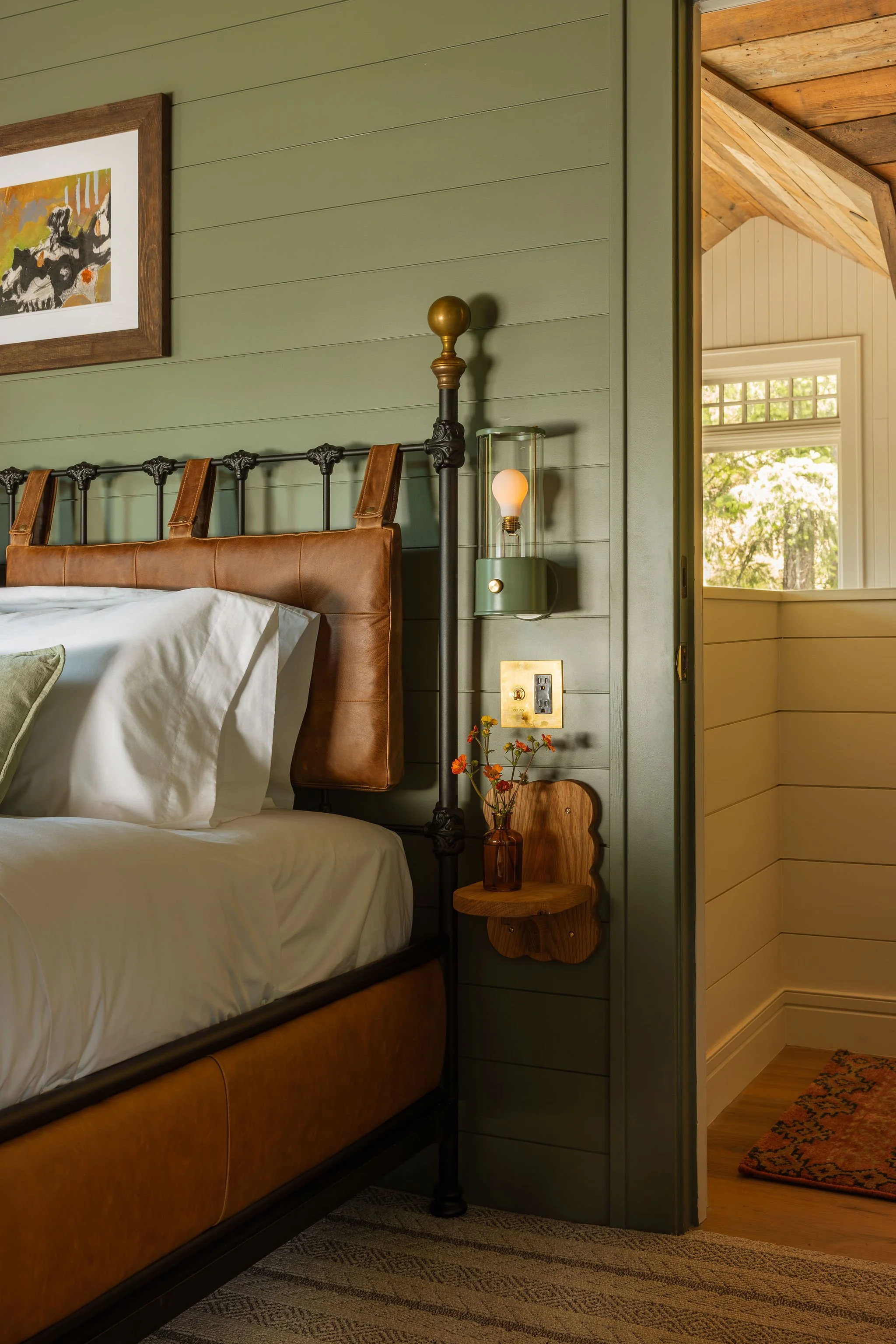 A bedroom with a green-shaded wall, featuring a black metal bed with a brown leather headboard and white bedding. There is a small wooden side table with a vase of orange flowers, a wall-mounted lamp with an exposed bulb, and a framed abstract artwork above the bed. A partially open door reveals a room with white paneled walls, a window with a view of trees, and a patterned rug.