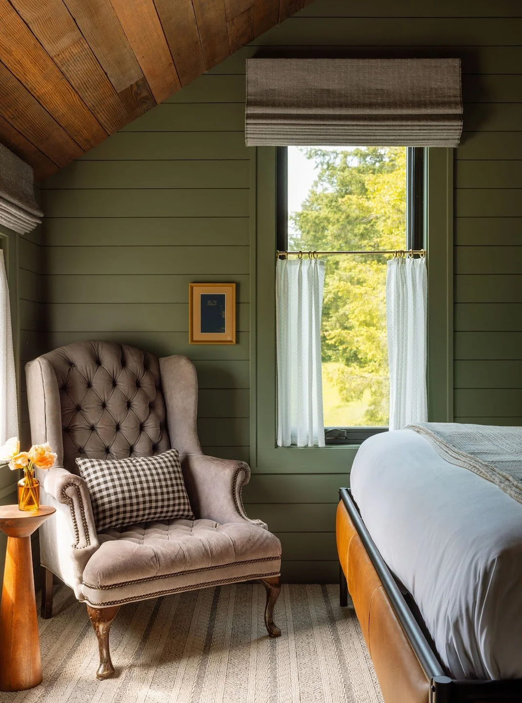 A bedroom corner with a tufted high-back armchair with a plaid pillow, a small wooden side table with a vase of flowers, a framed picture on a green shiplap wall, a window with white curtains and a view of green trees, and part of a bed with white bedding.