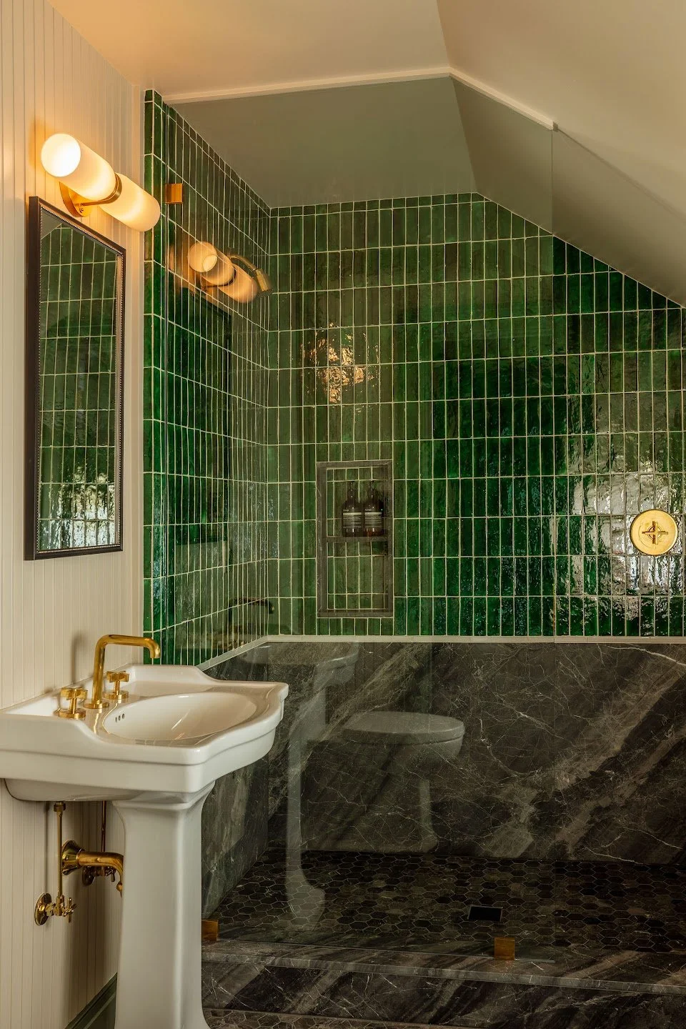 A bathroom with green tiled walls, dark marble floating vanity, and a white vintage-style pedestal sink with gold fixtures. Features include a mirror with gold trim, wall-mounted gold light fixtures, and a walk-in shower with similar green tiles and a recessed shelf.