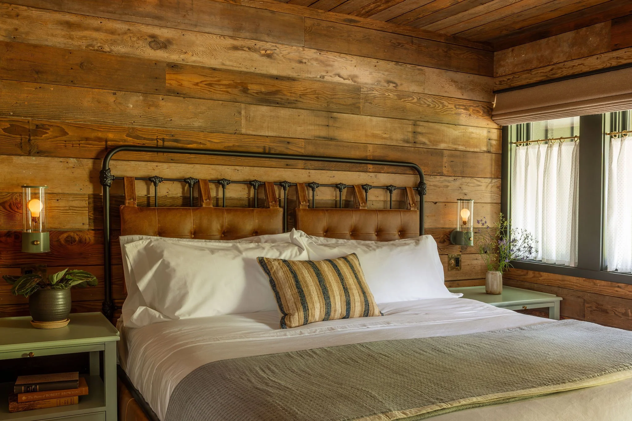 A cozy bedroom with a wooden wall and ceiling, a bed with white linens and decorative pillows, a small side table with a potted plant and books, a window with curtains, and warm ambient lighting from bedside lamps.