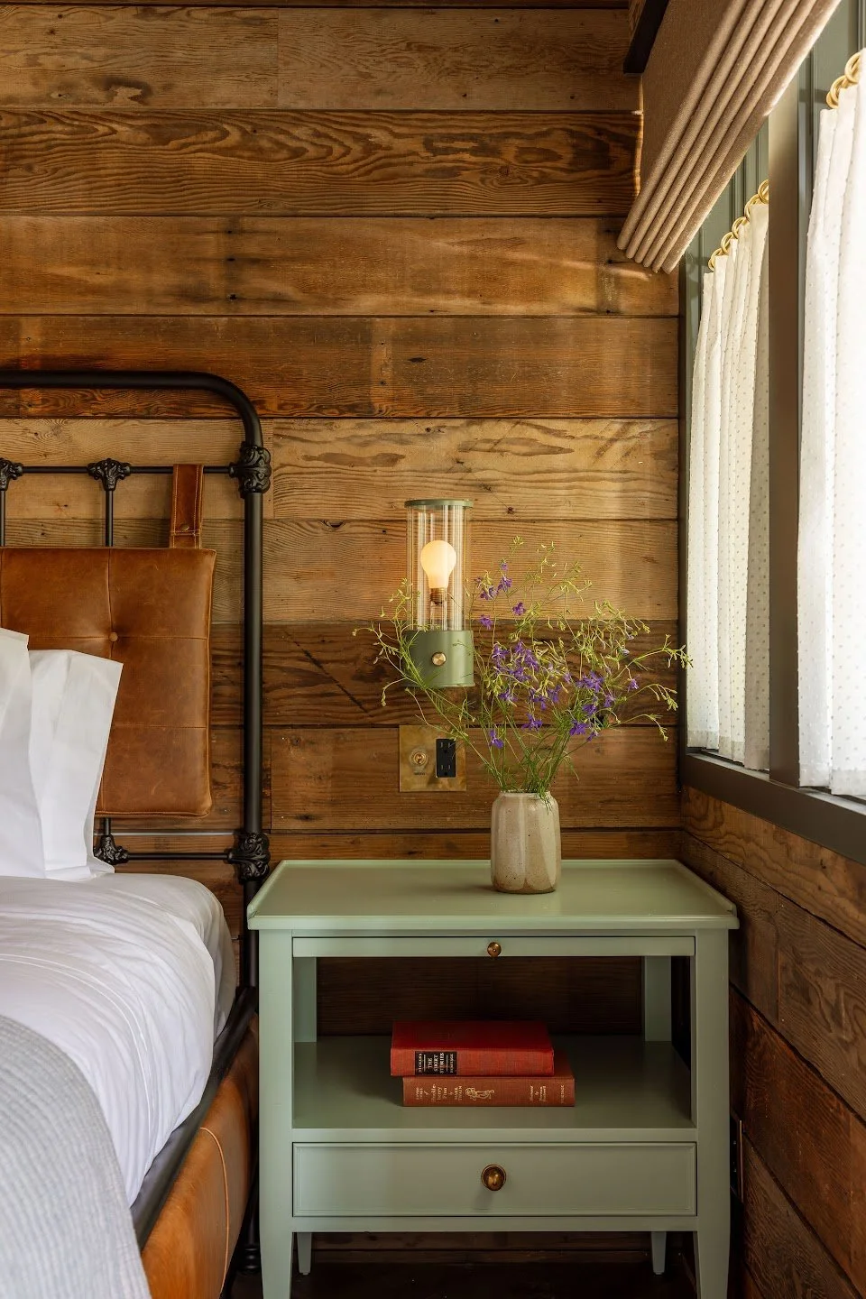 A bedroom bedside table with a vase of purple flowers, a wall sconce, and a window with curtains.
