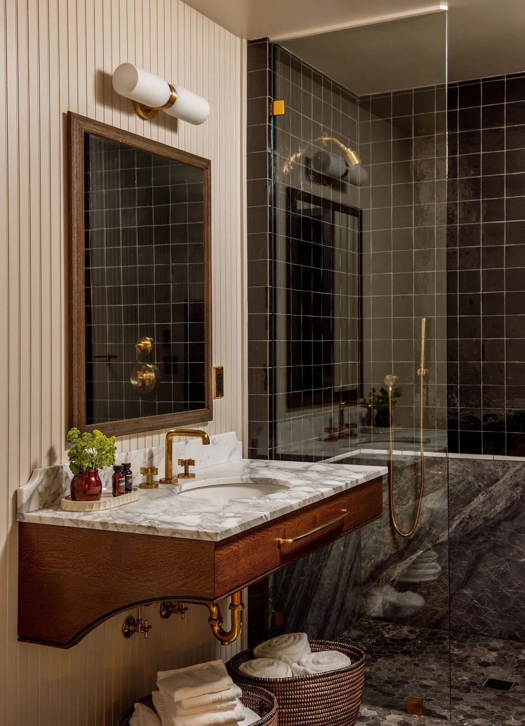 A luxurious bathroom with a marble sink, brass fixtures, a wooden framed mirror, a wall-mounted light, a plant, and a walk-in shower with dark tiled walls and gold fixtures.