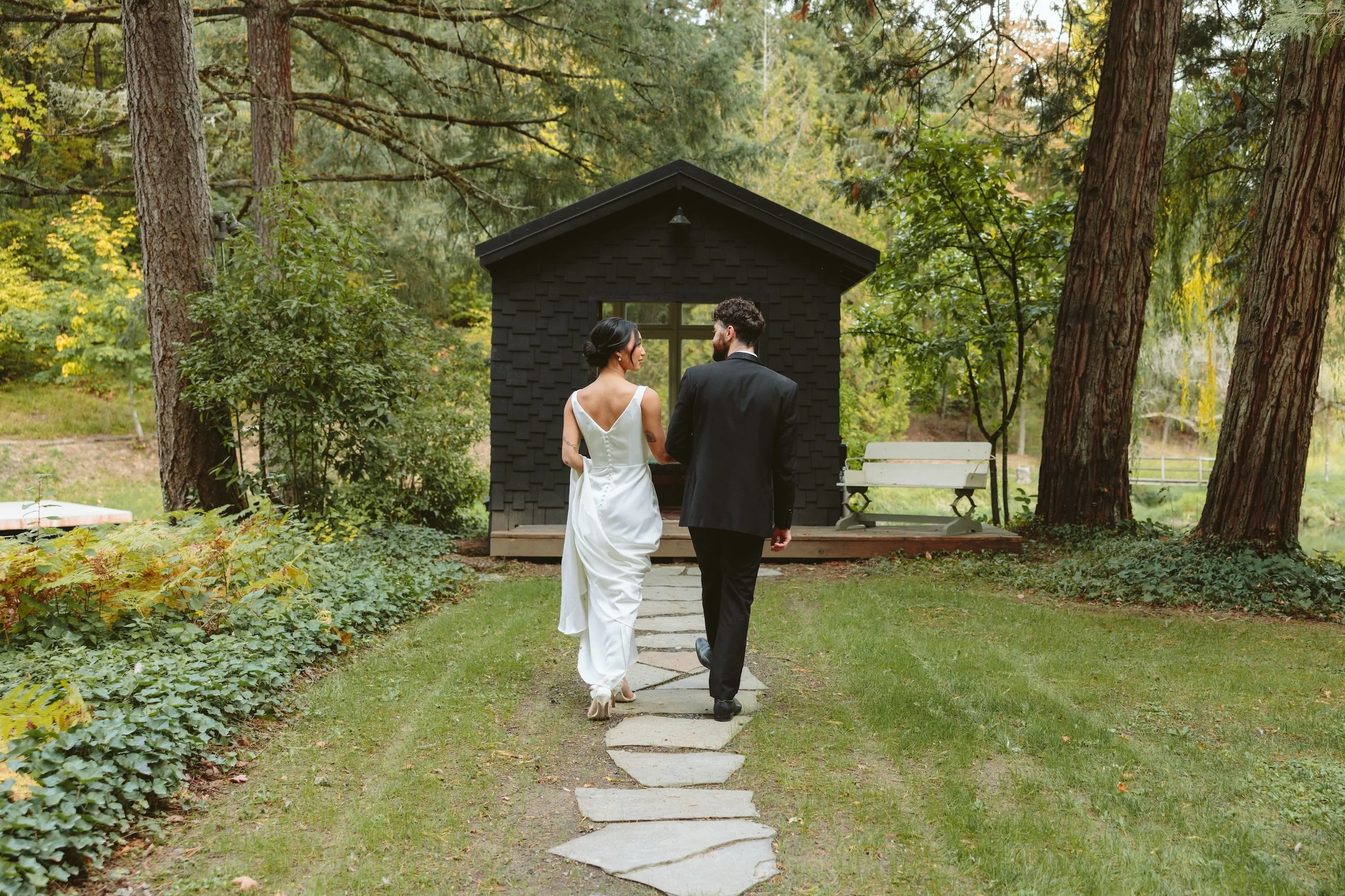 A bride and groom holding hands walking on stone stepping stones in a lush, green forest with tall trees and a small black wooden shed in the background.