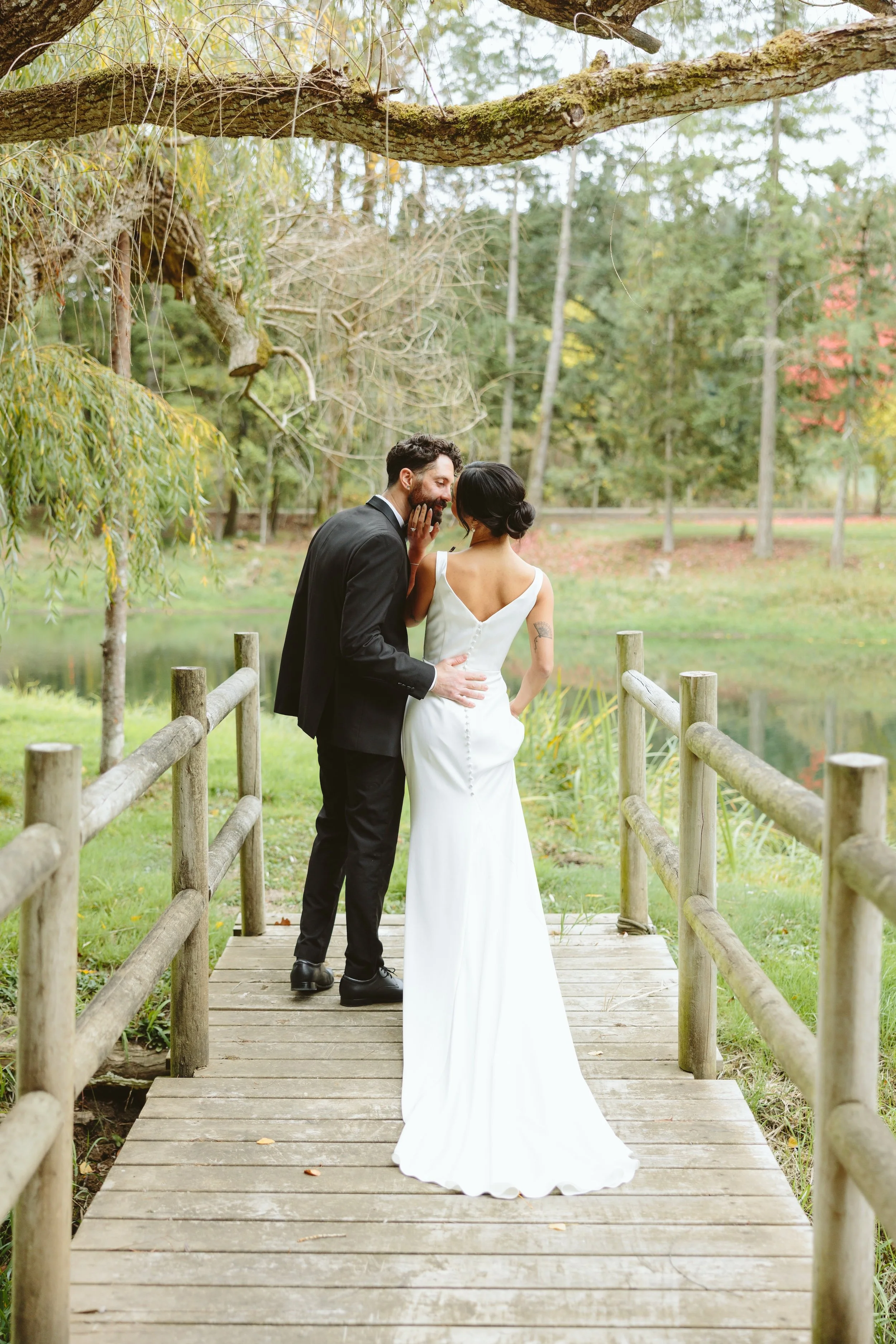 A bride and groom share a kiss on a wooden bridge in a park surrounded by trees.