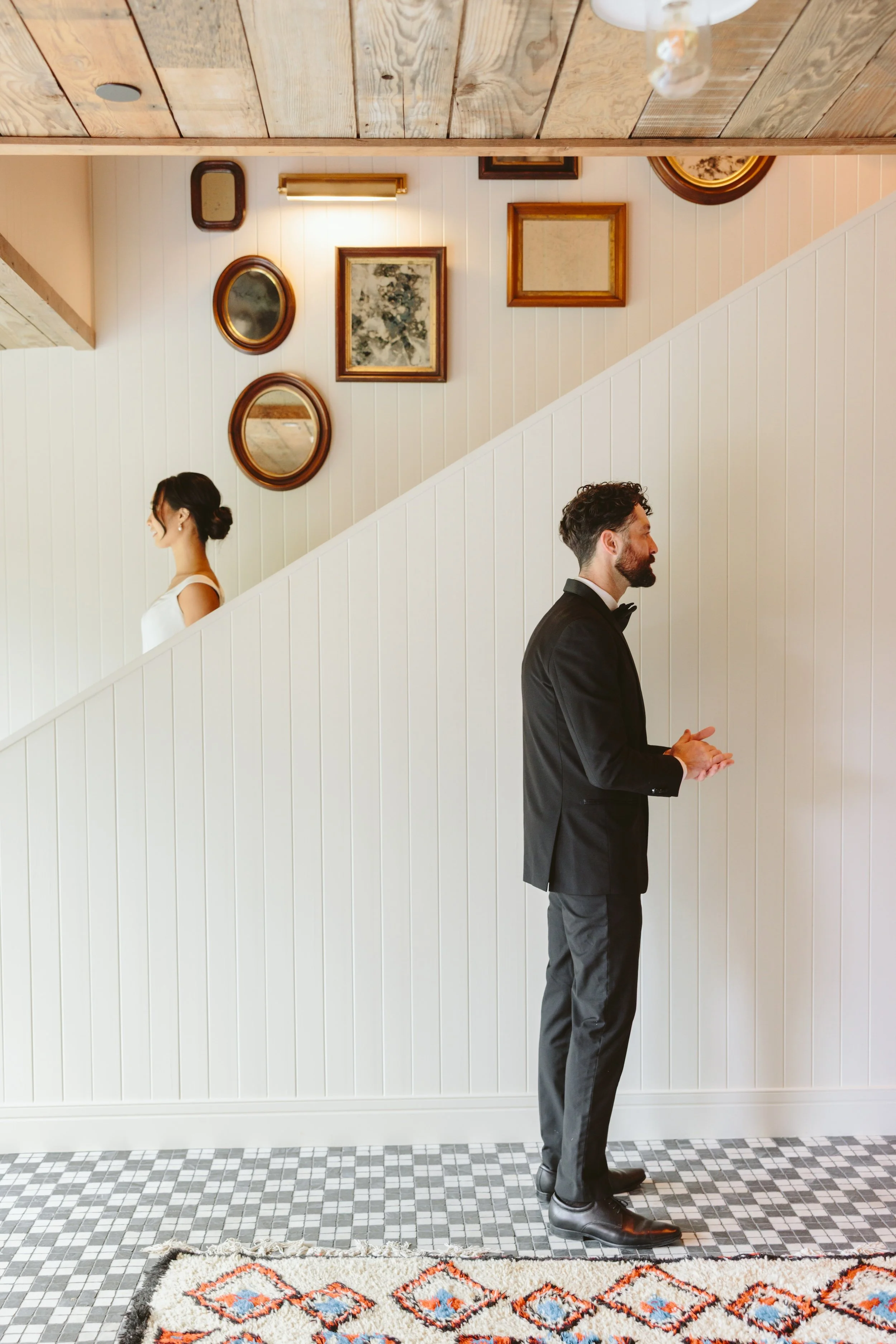 A man dressed in a black tuxedo with a bow tie stands with his hands clasped, looking to the right. A woman with dark hair in a bun, wearing a white dress, stands to the left with her profile visible. They are in a room with white wooden paneling on 