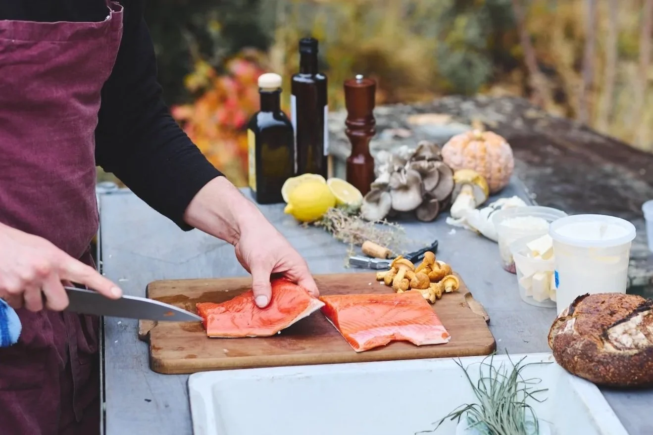 Person preparing salmon fillets on a wooden cutting board outdoors, surrounded by various ingredients and cooking supplies, with autumn foliage in the background.