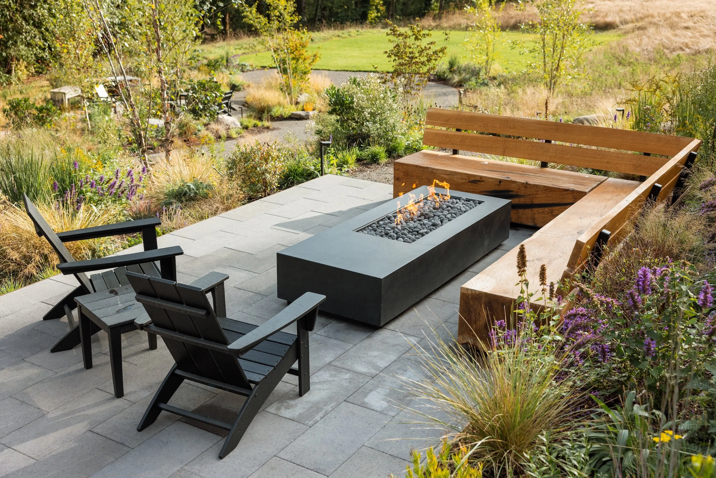 A backyard patio with black Adirondack chairs, a rectangular fire pit, and a built-in wooden bench, surrounded by lush garden plants and trees, with a grassy lawn in the background.