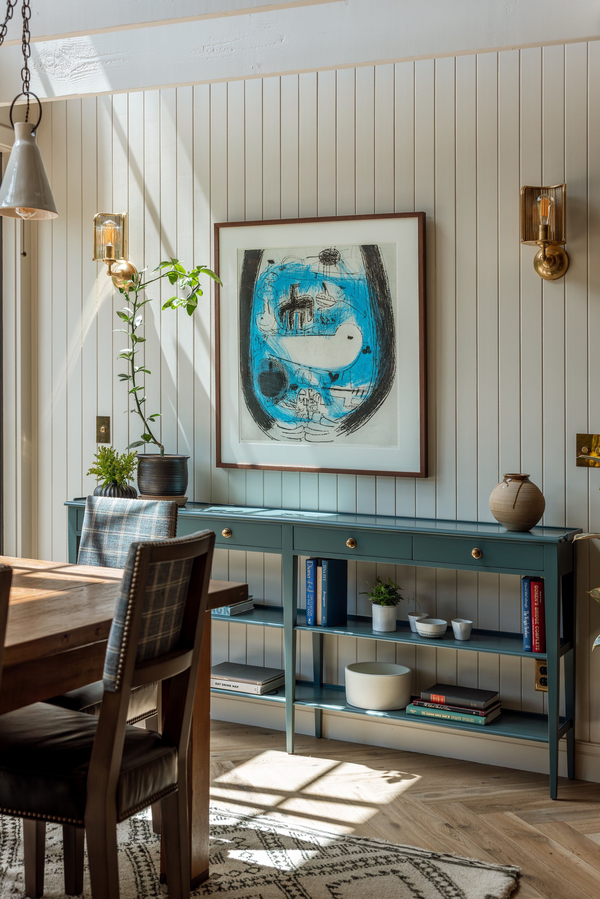Interior of a dining room with a wooden table, upholstered chairs, a blue bookshelf with books and decorative items, a framed abstract artwork, wall sconces, and a potted plant. Sunlight streams in through a window, casting shadows.