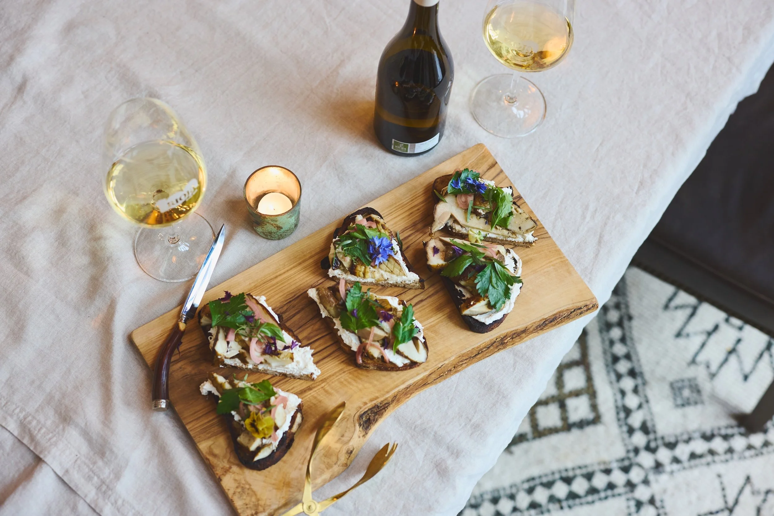 A wooden serving board with sliced sandwiches garnished with herbs and edible flowers, placed on a white tablecloth. Two glasses of white wine, a wine bottle, and a candle in a decorative holder are nearby.
