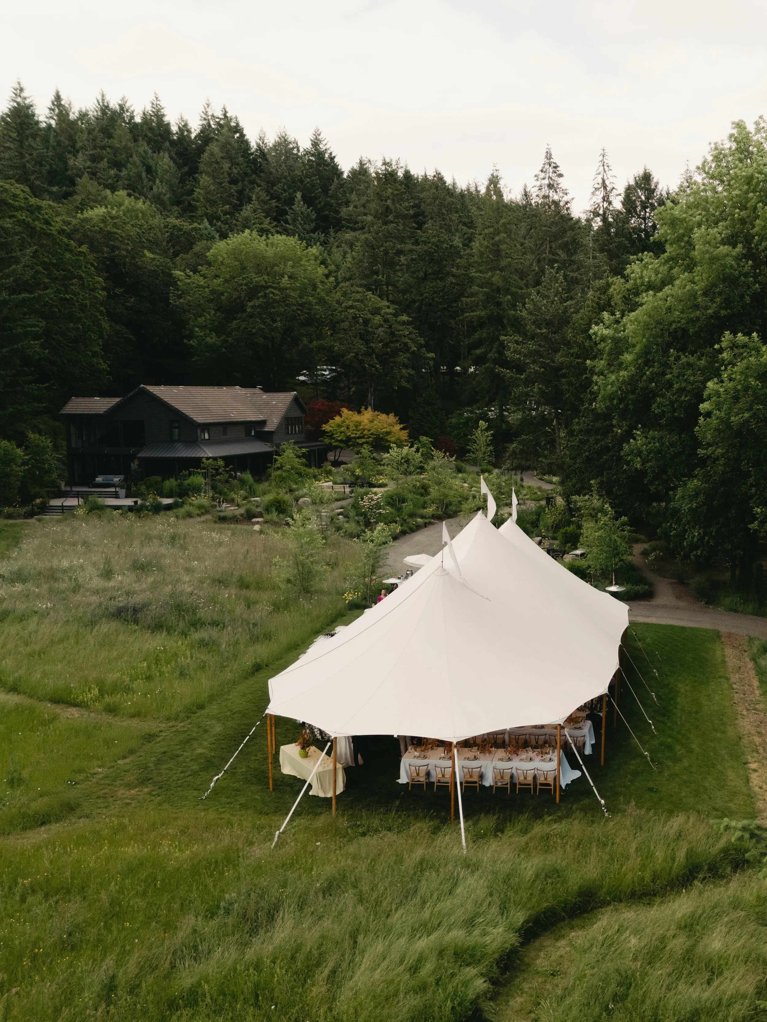 Outdoor wedding setup with a white tent and tables on a grassy area, surrounded by lush green trees and forest in the background.