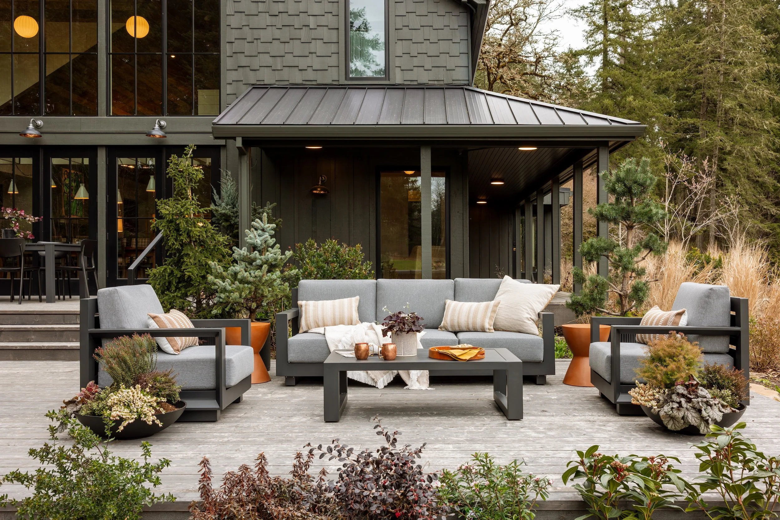 An outdoor patio area with gray cushioned benches and chairs, a black coffee table with decorative items, surrounded by potted plants and small trees, with a modern house in the background featuring large windows, a metal roofed overhang, and surroun