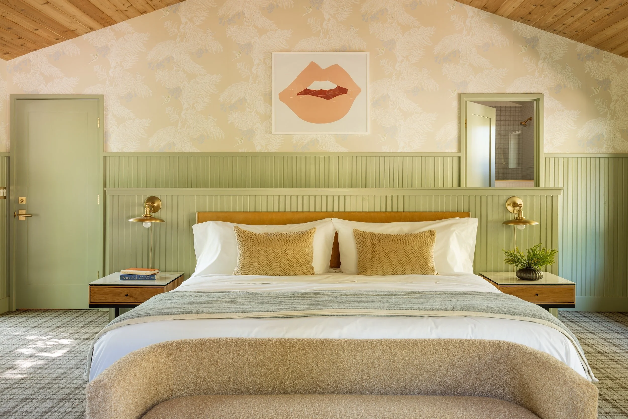 A cozy bedroom with a wooden vaulted ceiling, light green paneled walls, and a large bed with white bedding and beige pillows. There is a framed artwork of lips above the bed, and two gold bedside lamps. A windowed door leads to a bathroom with a sho