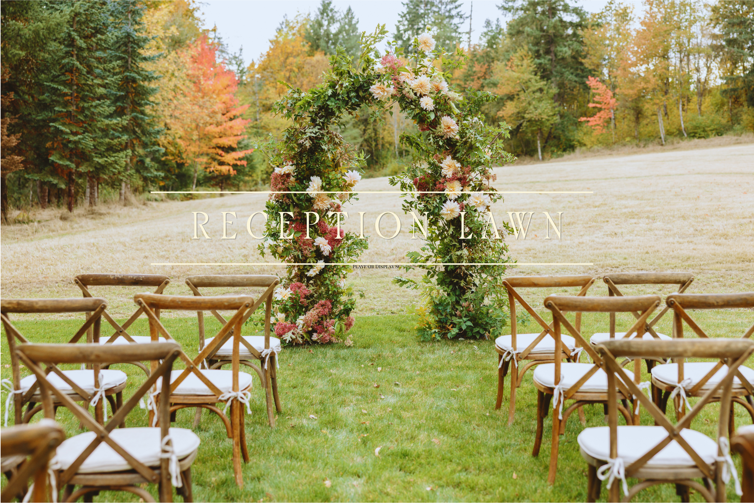 An outdoor wedding setup with rows of wooden chairs arranged on a grass lawn, facing a floral arch made of pink and white flowers and green foliage. In the background, there are trees with fall foliage in shades of green, orange, and red, and a partly cloudy sky.