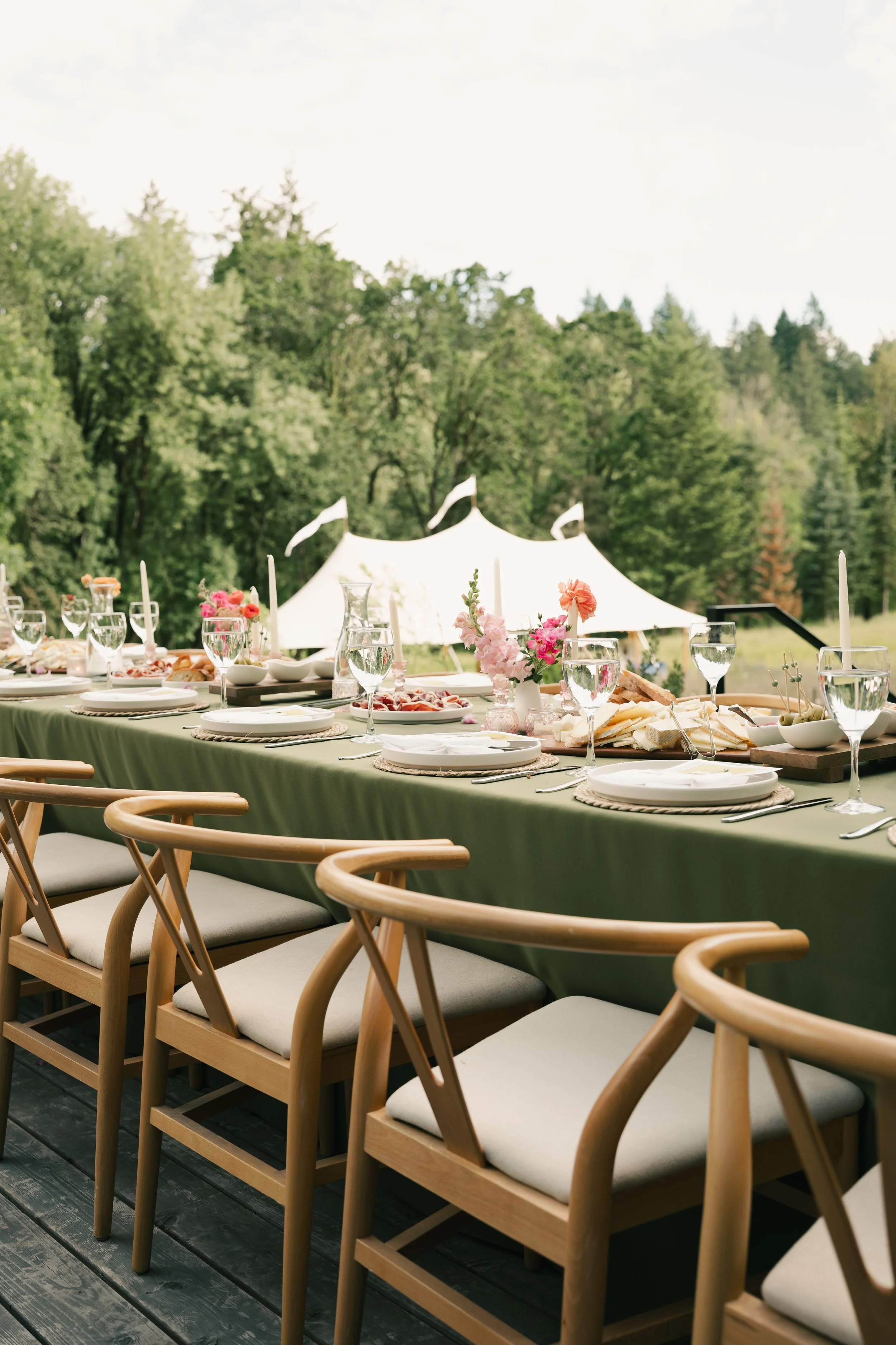 A long outdoor dining table with a green tablecloth, set with plates, wine glasses, and floral centerpieces. Wooden chairs with white cushions are arranged around the table. In the background, there is a white tent and lush green trees, indicating a 