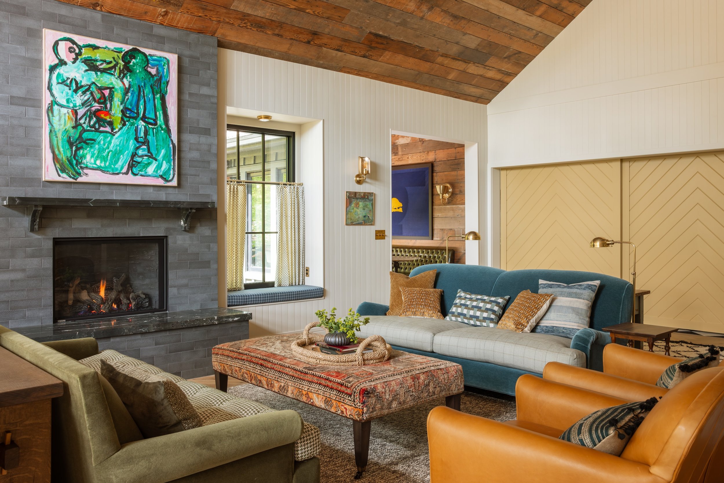 A cozy living room with a wood-paneled vaulted ceiling, a gray brick fireplace with a colorful abstract painting above it, a large window with yellow patterned curtains, a blue sofa with patterned pillows, a stone coffee table with plants, and leathe