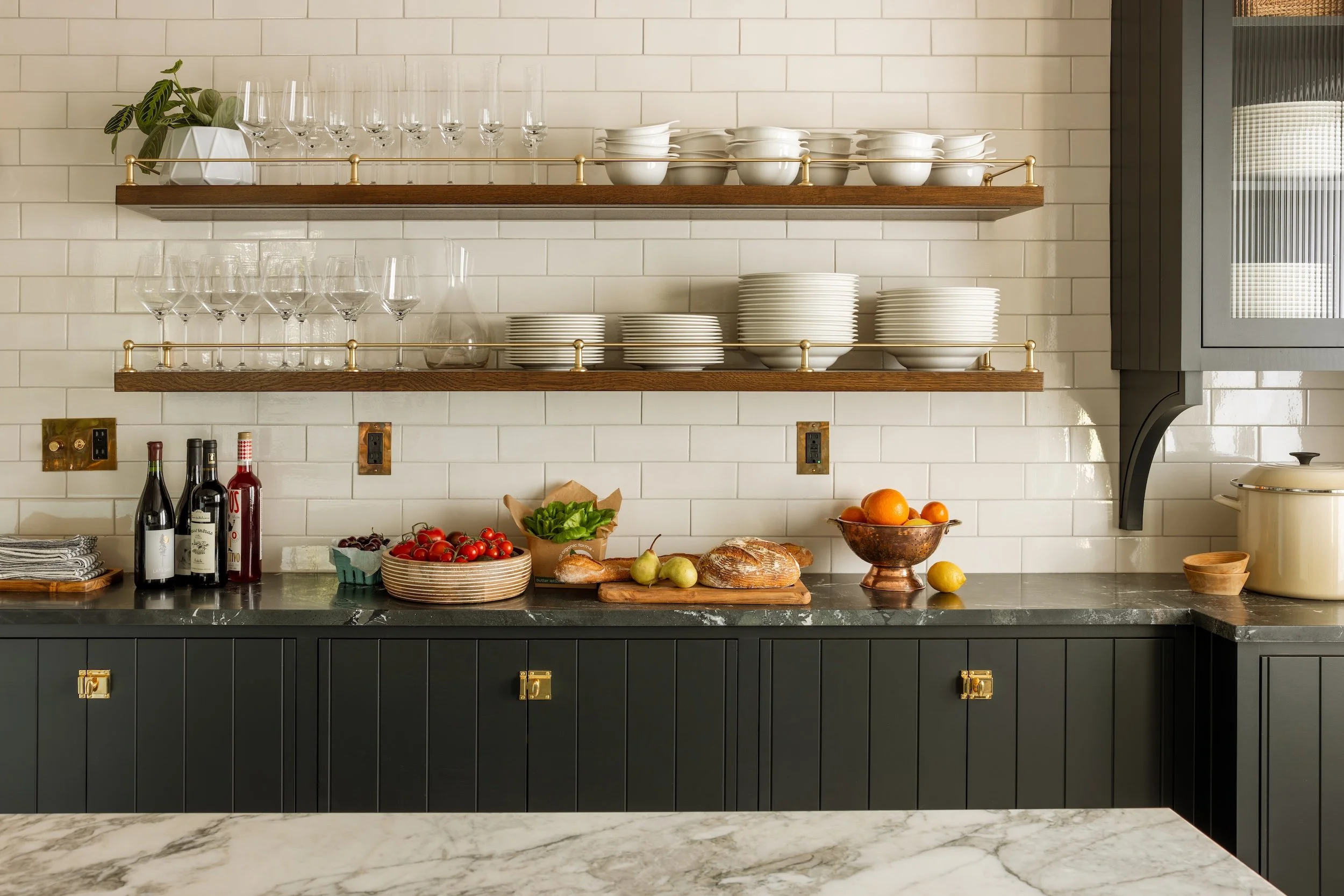 Kitchen shelves with glassware, dishes, and a plant, with a countertop below holding bottles, tomatoes, bread, and fruits, against a white tiled wall and dark cabinetry.