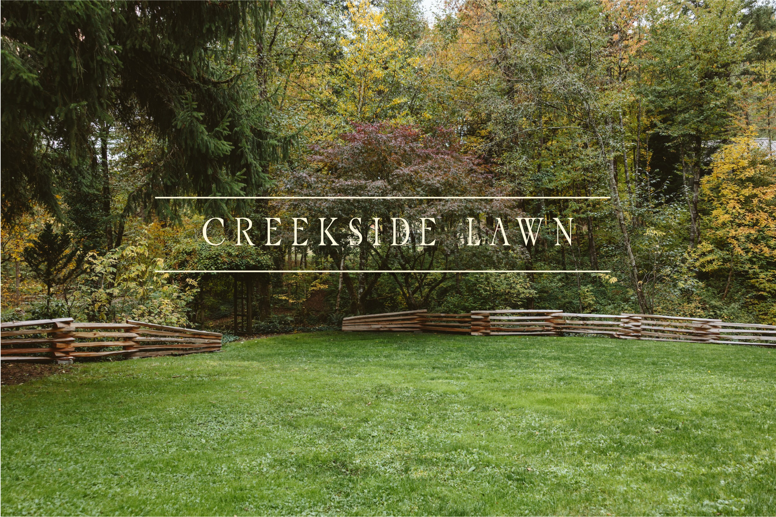 A lush green lawn with a wooden fence in front of a dense forest of trees with changing autumn foliage. The image features a sign or overlay that reads 'Creekside Lawn'.