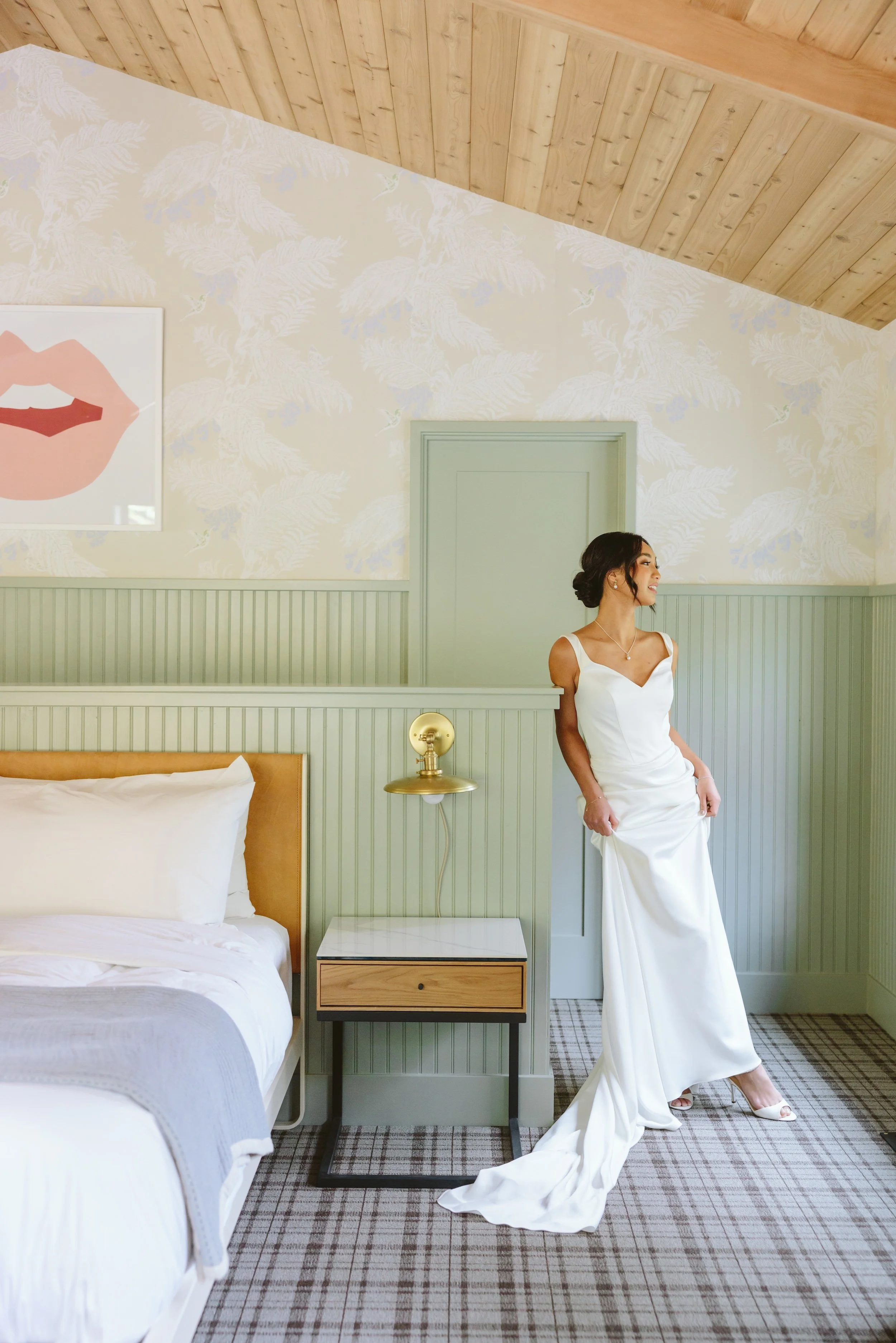 A woman in a white dress is sitting beside a bed in a hotel room with green paneled walls, a wooden bedside table, and a sky-patterned wallpaper. She is smiling and looking to her left.