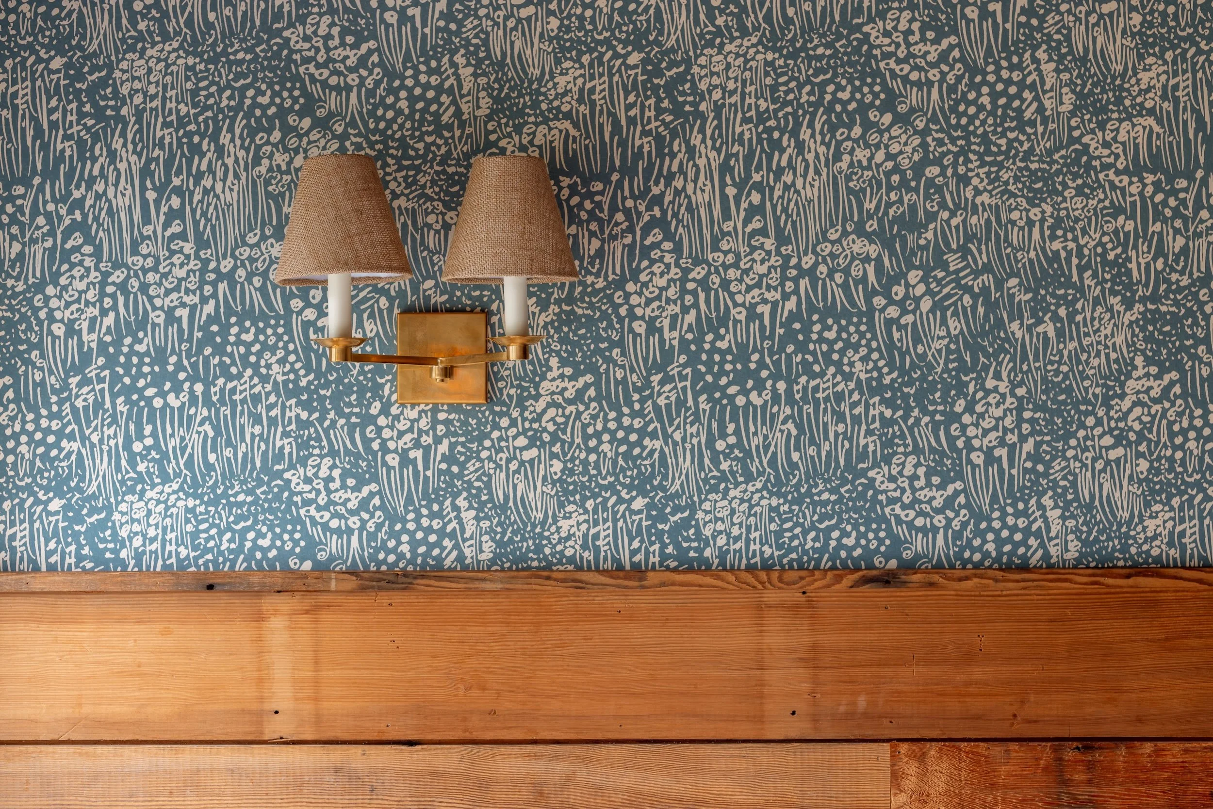 A wall with a dark blue background and a white abstract floral pattern, with two wall-mounted lamps with beige shades and a brass fixture above a wooden surface.
