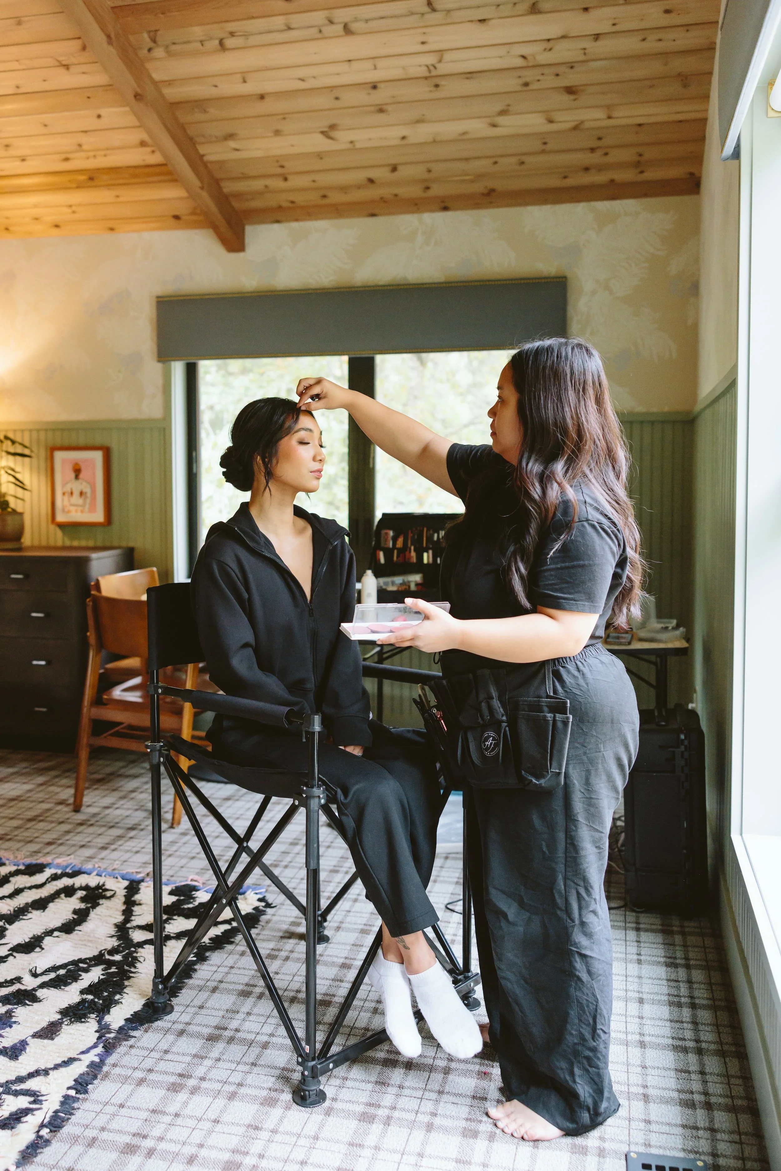 A makeup artist applies makeup to a woman sitting in a chair. The woman has dark hair styled in an updo and is wearing a black jacket. The artist has long, wavy brown hair and is holding a makeup palette. They are in a cozy room with wooden ceiling b