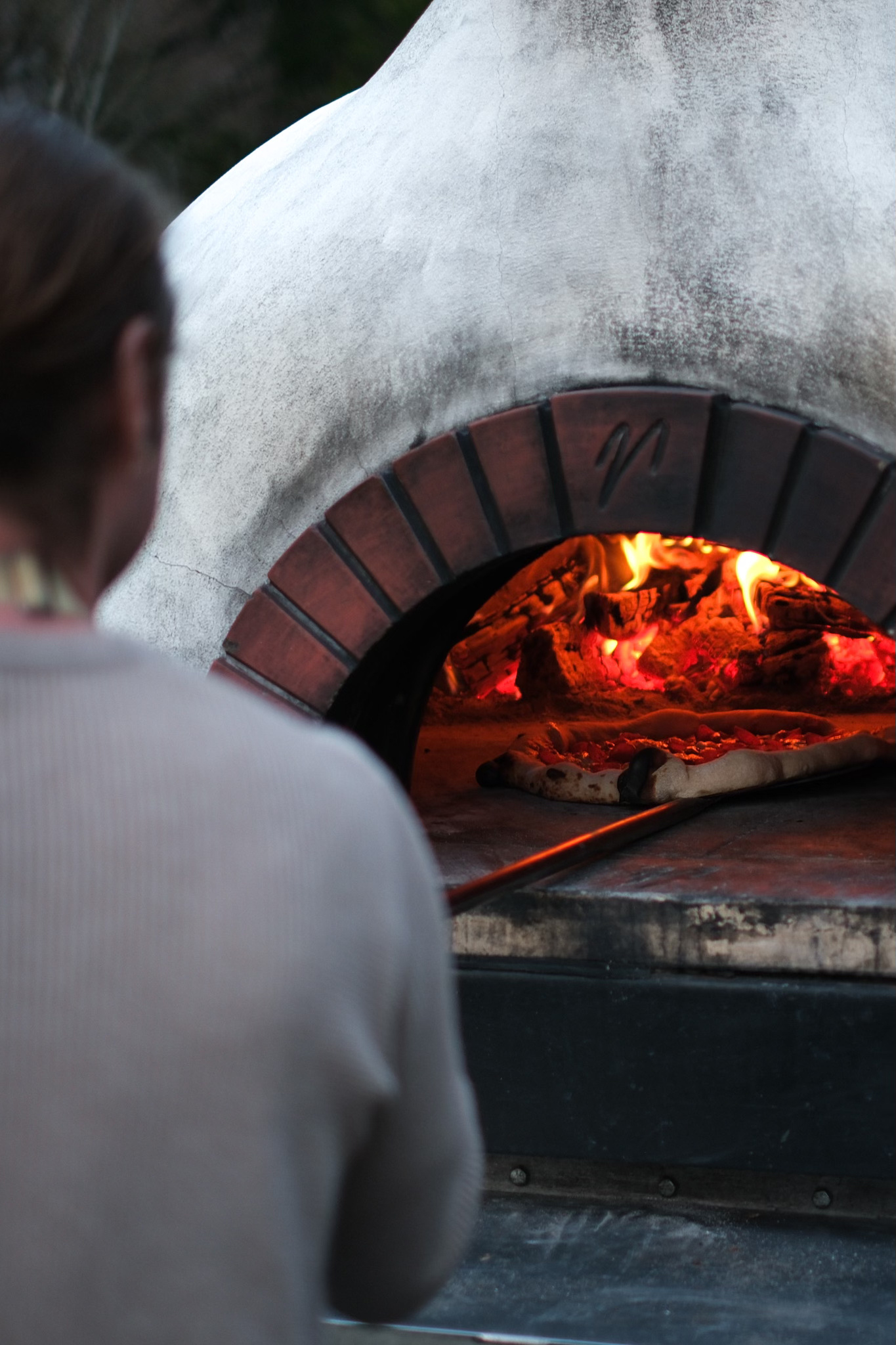 A person with brown hair and a light-colored top watching a pizza bake in a wood-fired oven with glowing fire and flames.