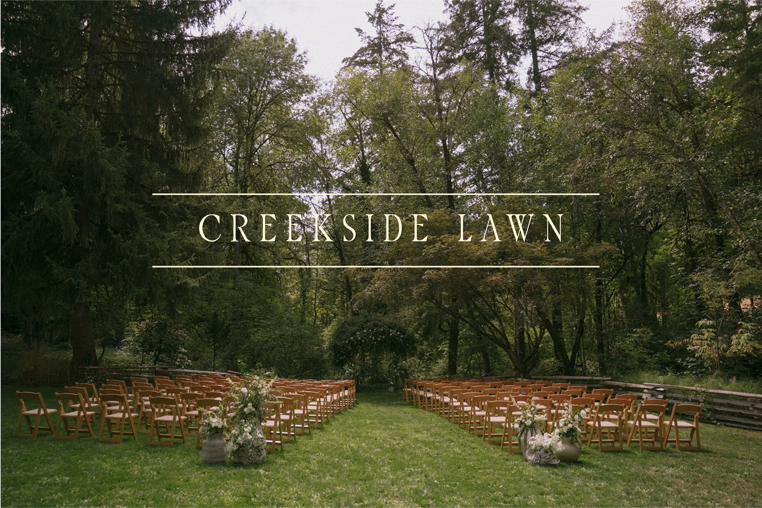 A lush green lawn with a wooden fence in front of a dense forest of trees with changing autumn foliage. The image features a sign or overlay that reads 'Creekside Lawn'.
