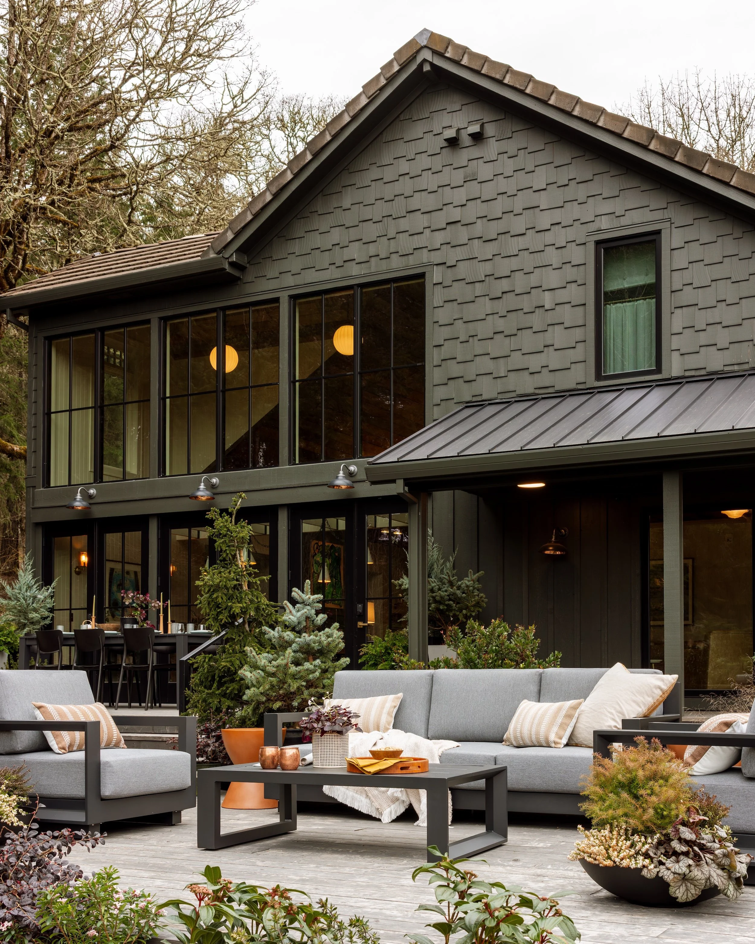 A modern outdoor patio area with gray cushioned seating, decorative pillows, a coffee table with plants and decorative items, and an enclosed sunroom with large glass windows and green plants outside. The patio has a wooden deck and surrounded by gre
