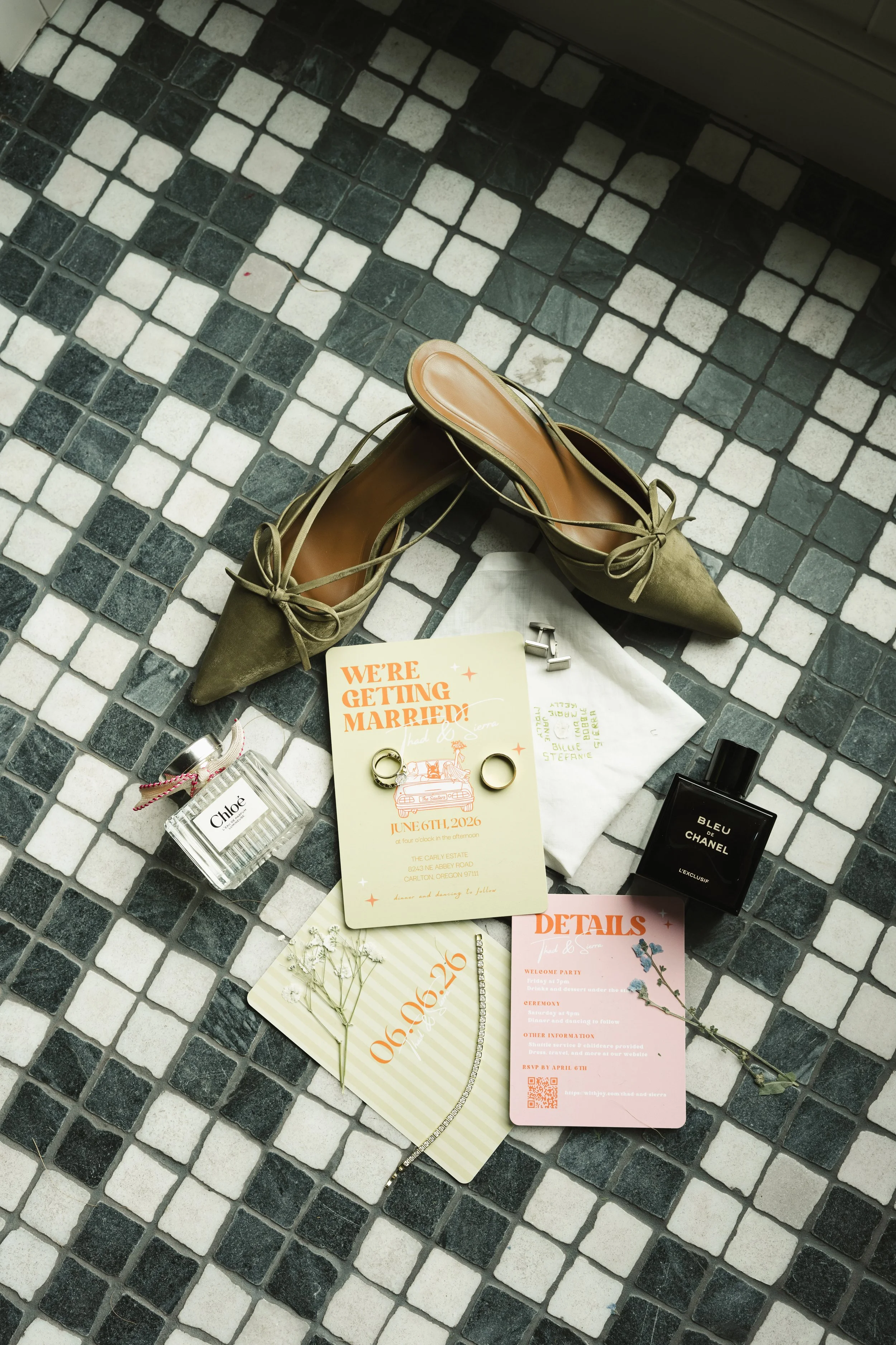 A pair of olive green pointed toe flats shoes with ankle straps and bows, placed on a tiled floor. Surrounding items include perfume bottles, wedding invitations, and small accessories, indicating a casual or preparatory setting.