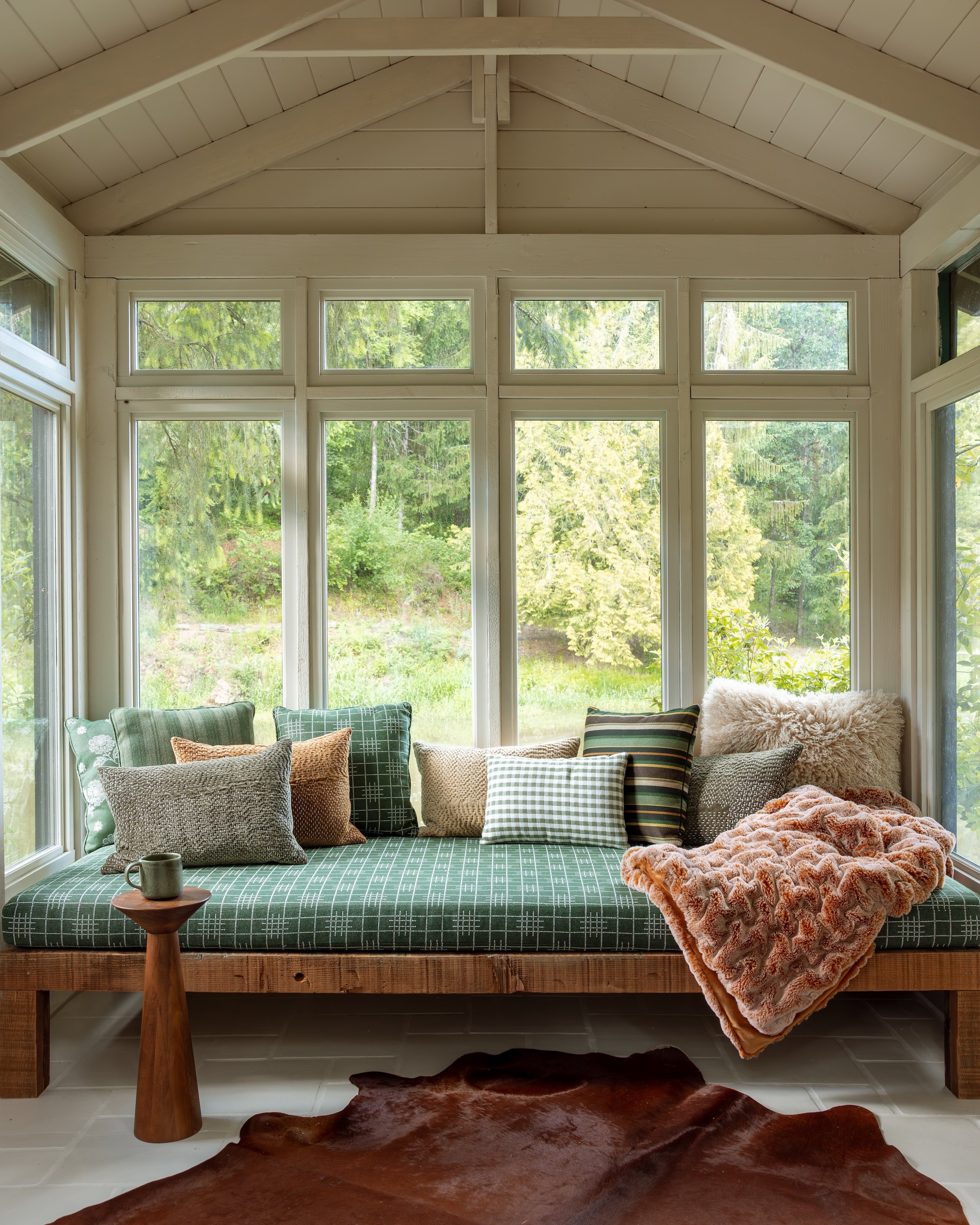 A cozy sunroom with large windows showing a lush green landscape outside. The room features a built-in bench with green checkered fabric cushions, a variety of throw pillows, and a plush blanket. A round wooden side table holds a mug, and a brown ani