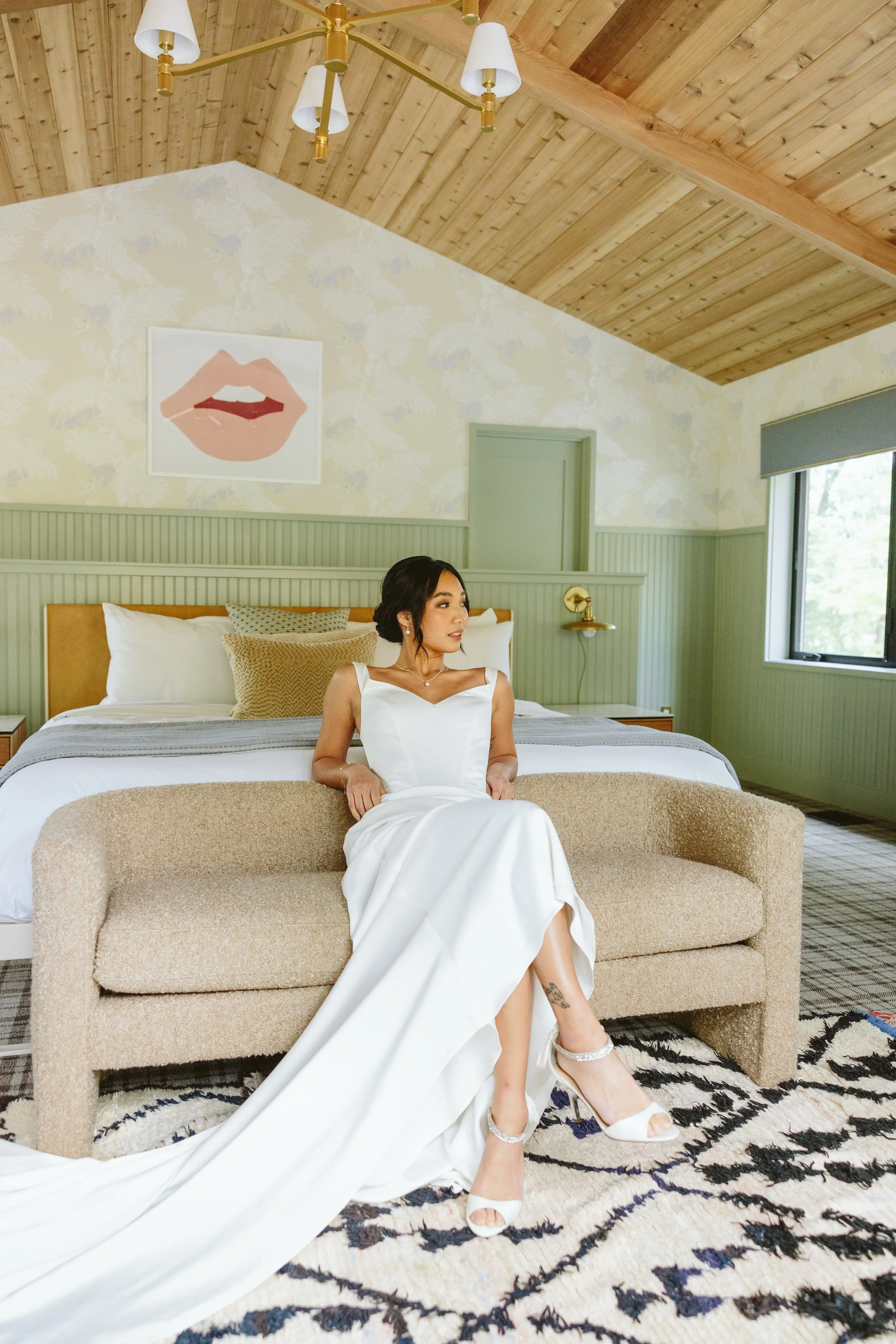 Woman wearing a white dress sitting on a beige sofa in a bedroom with wooden ceiling, green paneling, artwork of lips on wall, and a large window.