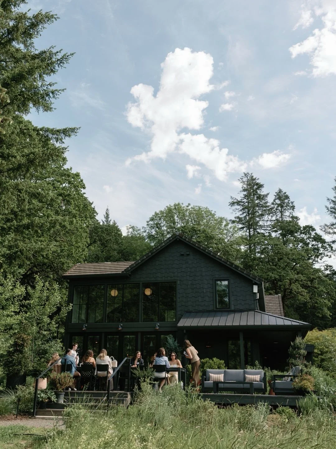 A group of people gathered on a patio outside a black house with large windows, surrounded by lush greenery and tall trees, under a partly cloudy sky.