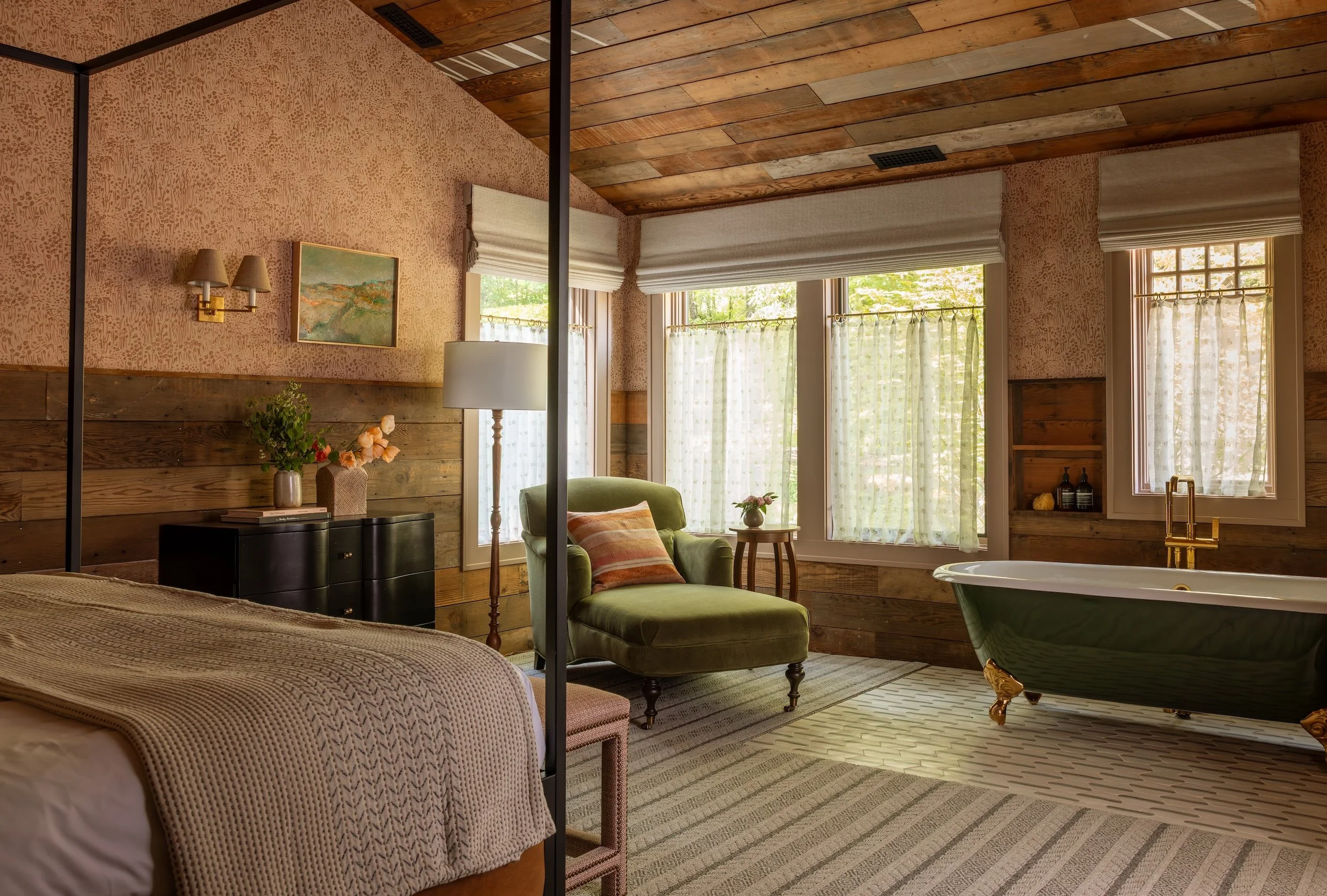 A cozy bedroom with a rustic design featuring a bed with textured bedding, a green armchair beside a small wooden table with flowers, framed artwork on textured pink wallpaper, natural light from large windows with white curtains, and wooden accents 