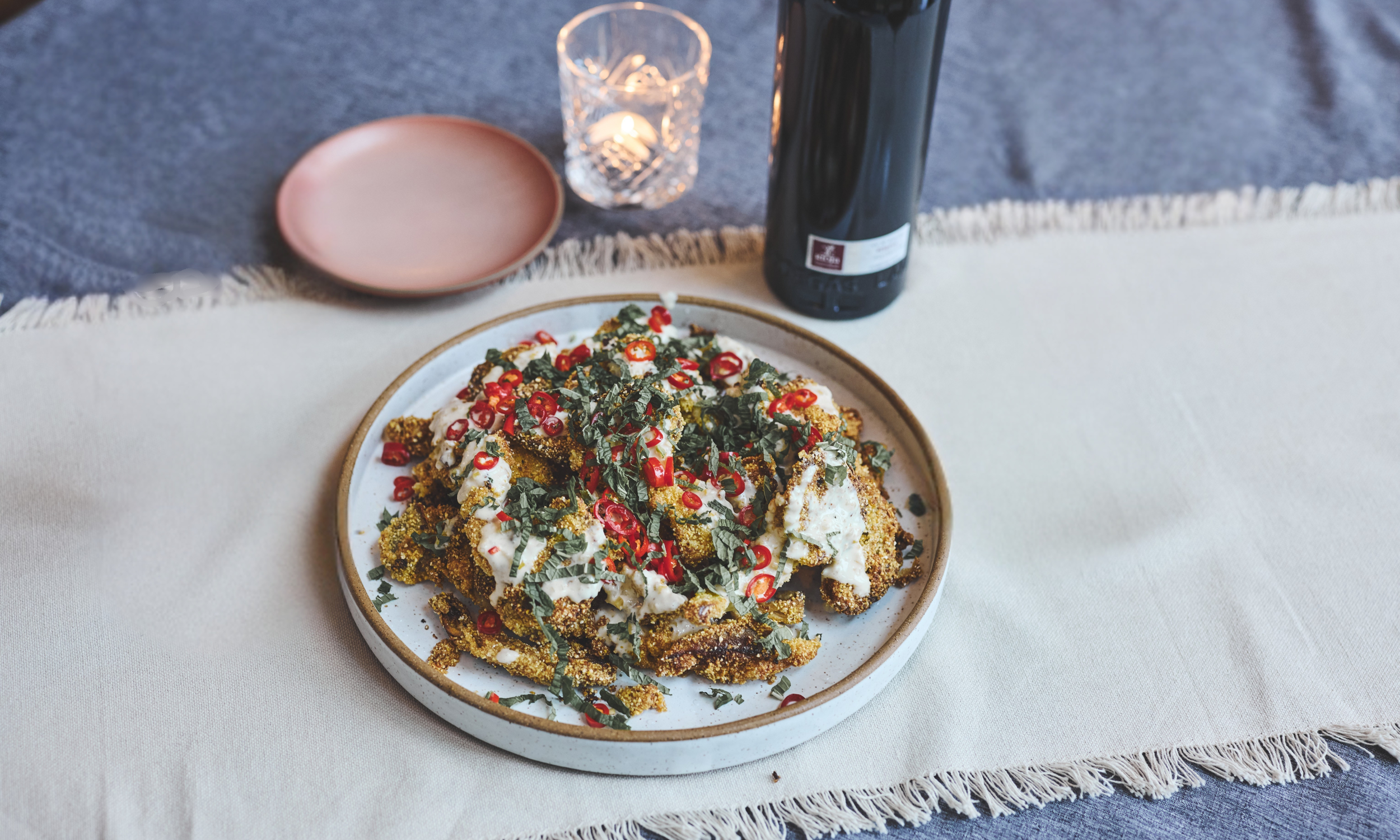 A plate of fried chicken topped with chopped red chili peppers, herbs, and a creamy sauce, set on a white tablecloth with a pink plate, a glass candle holder with a lit candle, and a dark bottle in the background.
