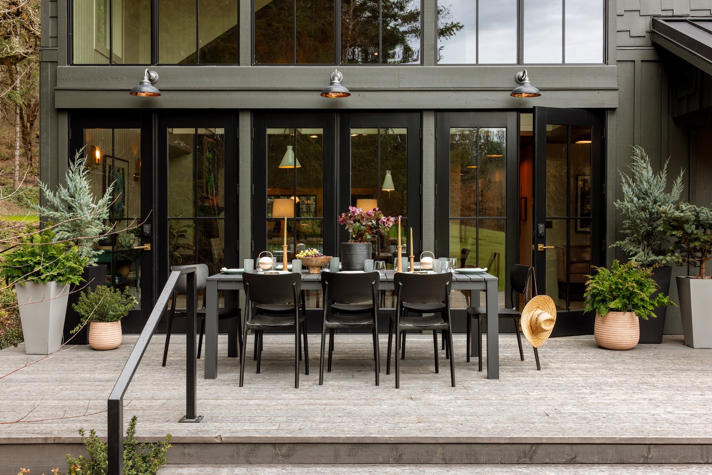A modern house with large glass doors and windows, outdoor patio with a dining table set for a meal, surrounded by potted plants and trees.