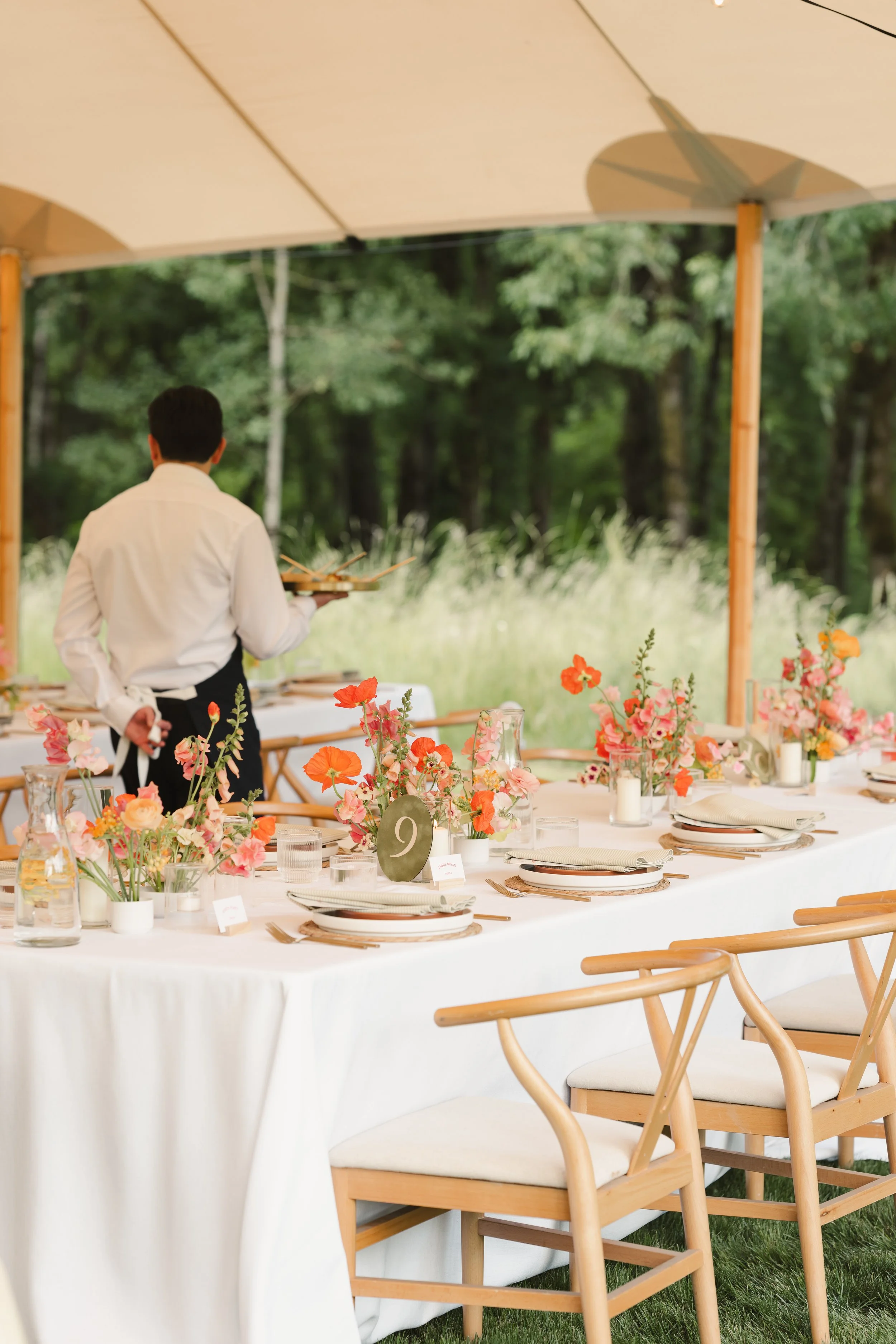 An outdoor wedding reception setup with a long table decorated with pink and orange flowers, glassware, and place settings under a large beige umbrella. Wooden chairs are arranged around the table, and a person in a white shirt and black apron is standing nearby with his back to the camera, holding a tray. The background features trees and greenery.