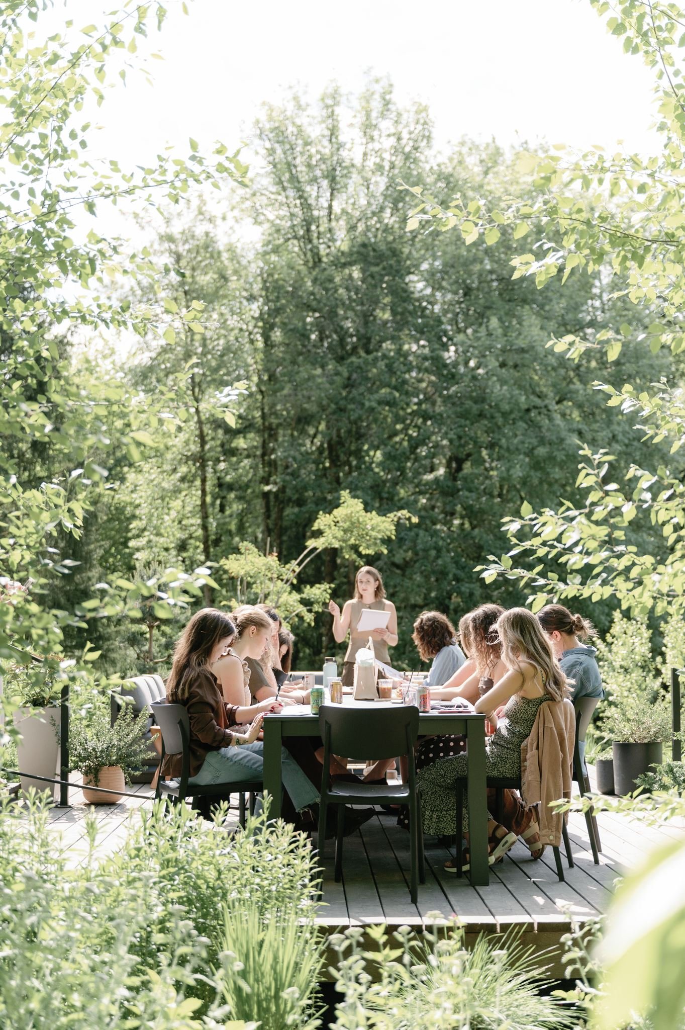 Group of people seated at outdoor dining table surrounded by green plants and trees, with a woman standing and speaking to the group in a garden setting.