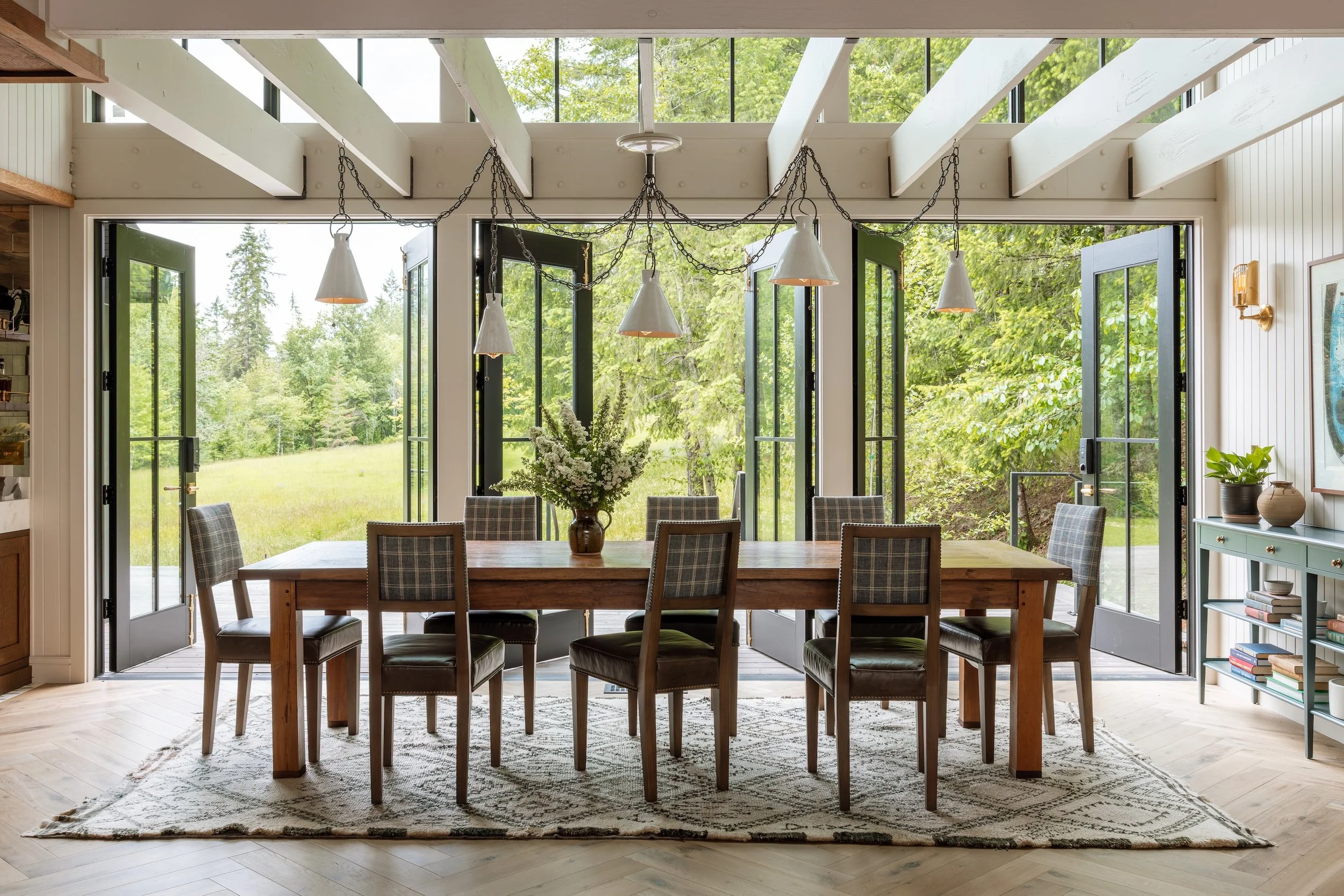 Dining room with a large wooden table, surrounded by seven plaid upholstered chairs, in front of large open glass doors leading to a green outdoor landscape. A vase with white flowers is on the table. Pendant lights with chain suspensions hang above 