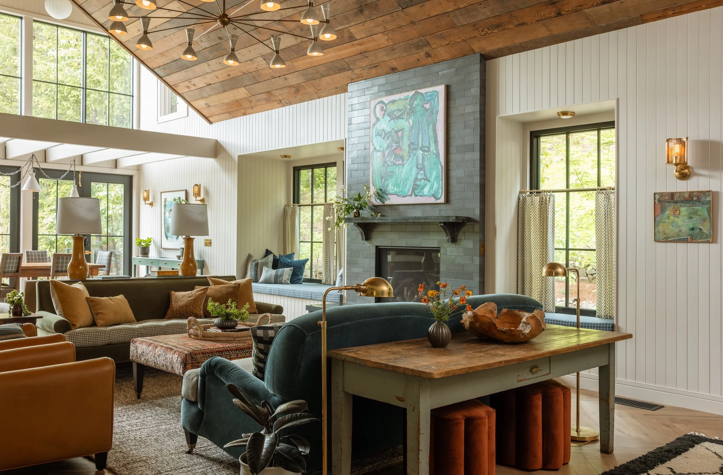 A spacious living room with a high, wood-paneled ceiling and large windows letting in natural light. The room features a stone fireplace with a colorful abstract painting above it, and various seating options including sofas and chairs with pillows. 