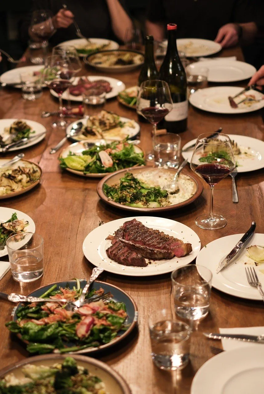A dinner table filled with various dishes including salad, steak, and other plated foods, along with glasses of red wine and bottles of wine.