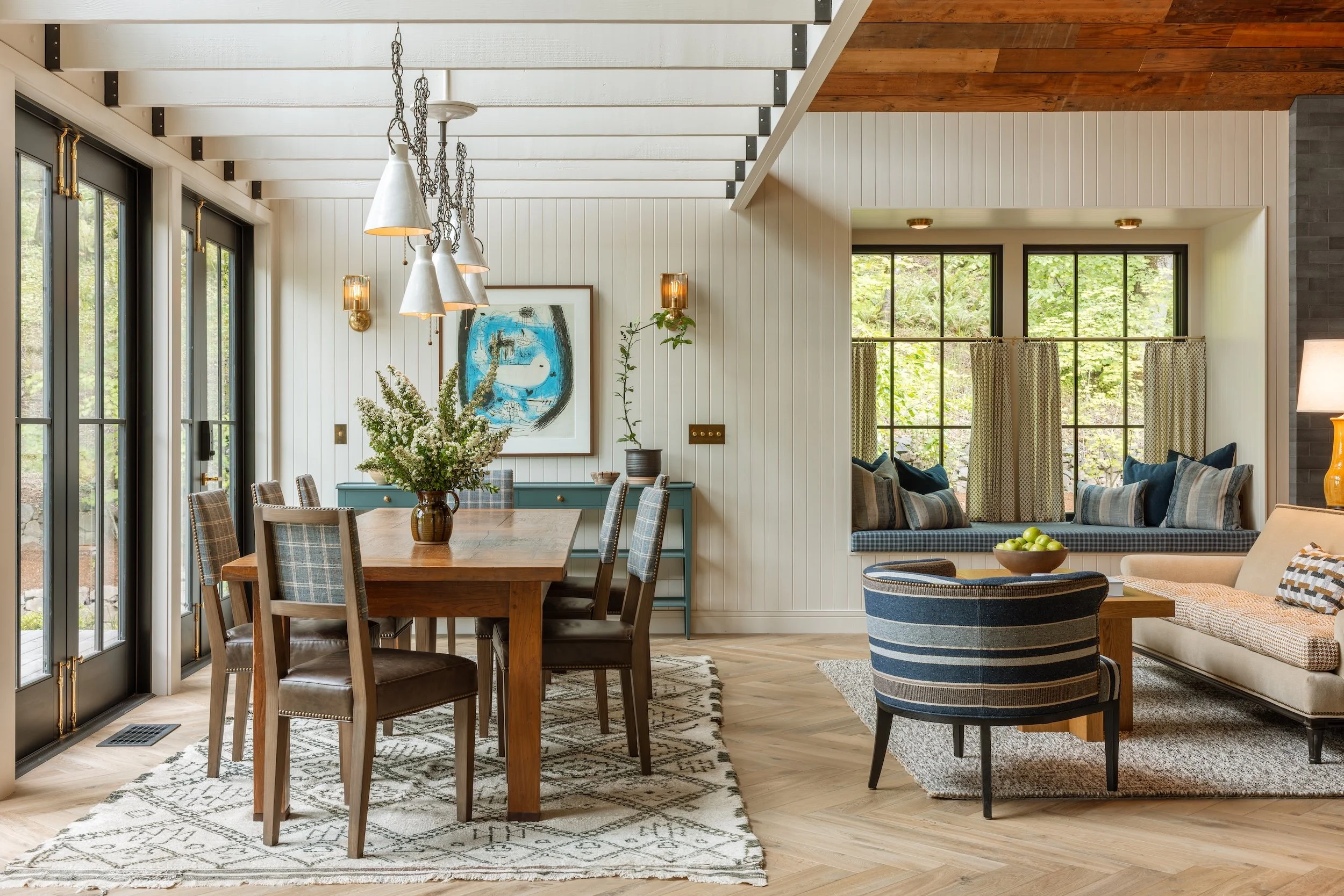 Interior view of a modern dining and living area with large windows, wooden floors, a dining table with plaid chairs, a blue sideboard with plants and artwork, a round striped armchair, and a beige sofa with cushions.