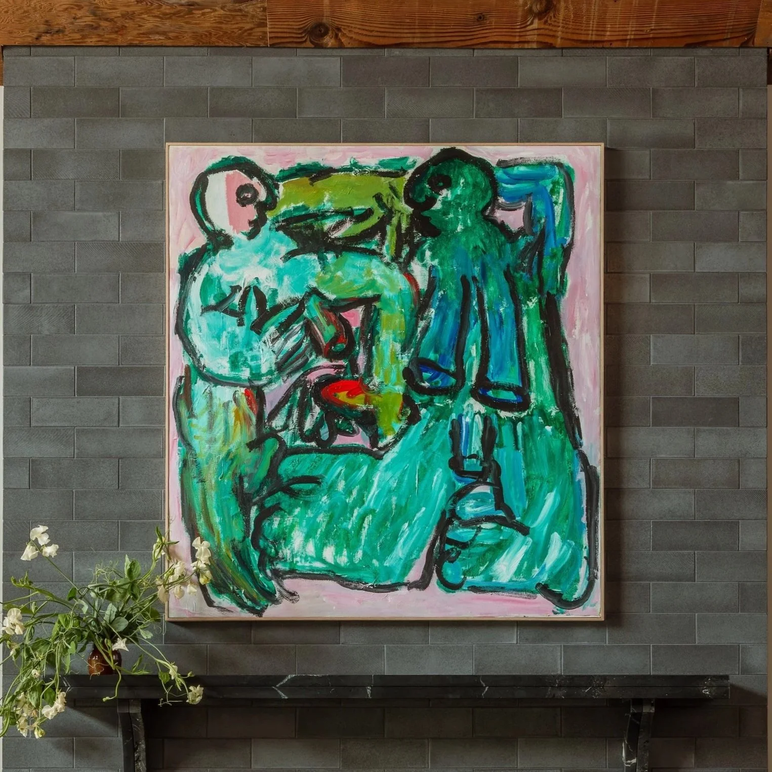 Abstract painting of two figures, one sitting and one standing, created with bold black brushstrokes and bright colors including green, pink, blue, and red, on a framed canvas displayed on a gray brick wall.