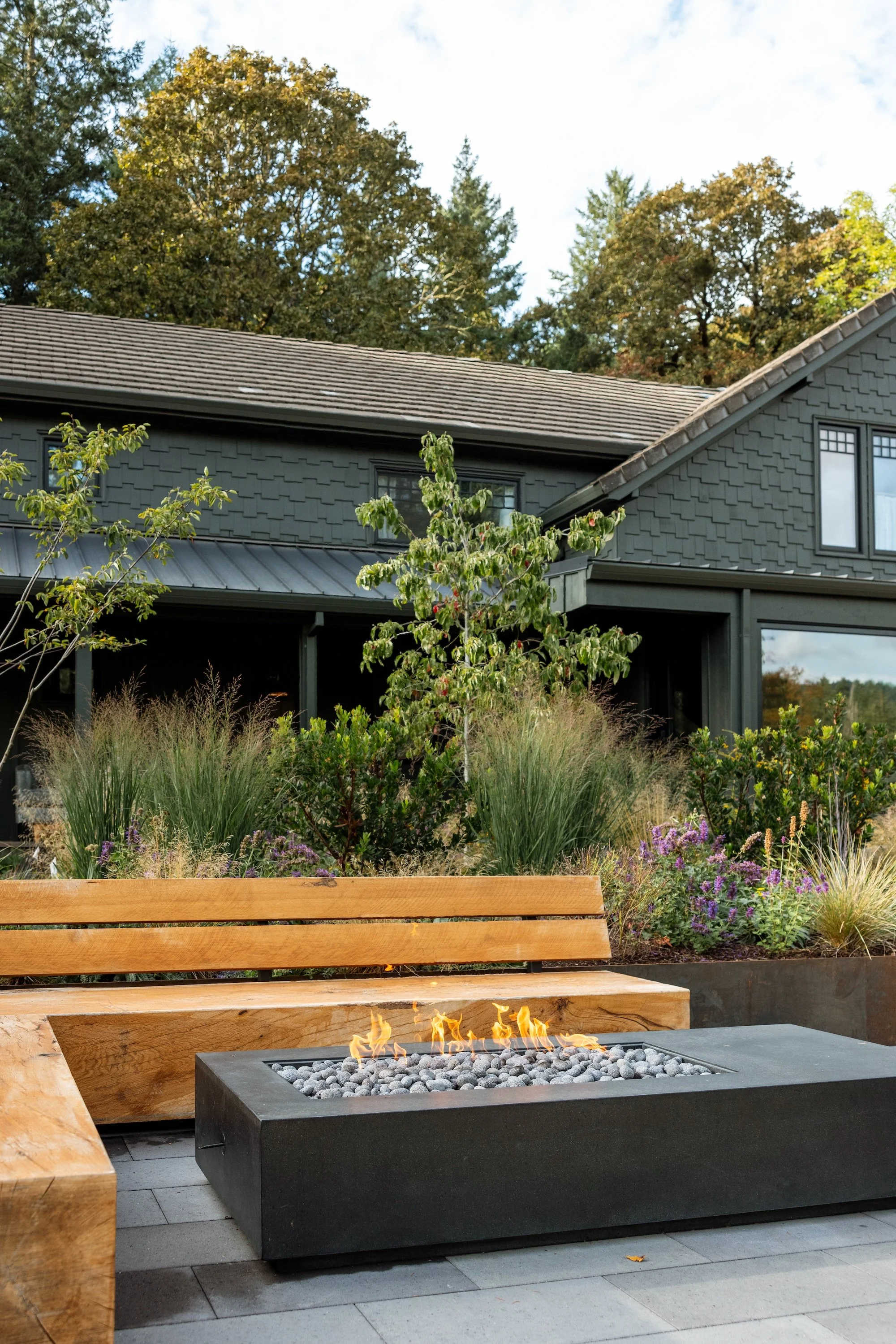 An outdoor patio area featuring a modern rectangular fire pit with flames, a wooden bench, lush garden plants, and a house with dark gray siding and a shingled roof, with trees in the background.