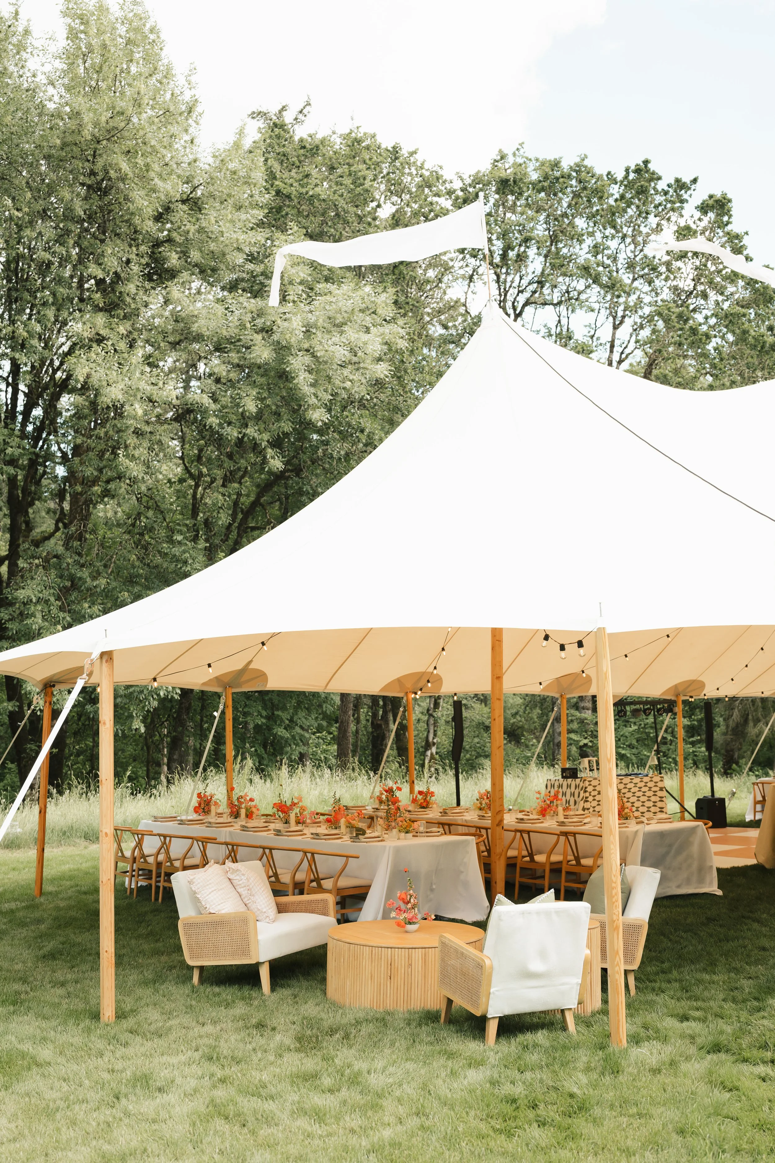 A large outdoor event tent with white fabric and wooden support poles, set up in a grassy area with trees in the background. The tent has string lights hanging underneath and a long dining table decorated with flowers. There are also white lounge chairs and a small round table with a flower arrangement in the foreground.