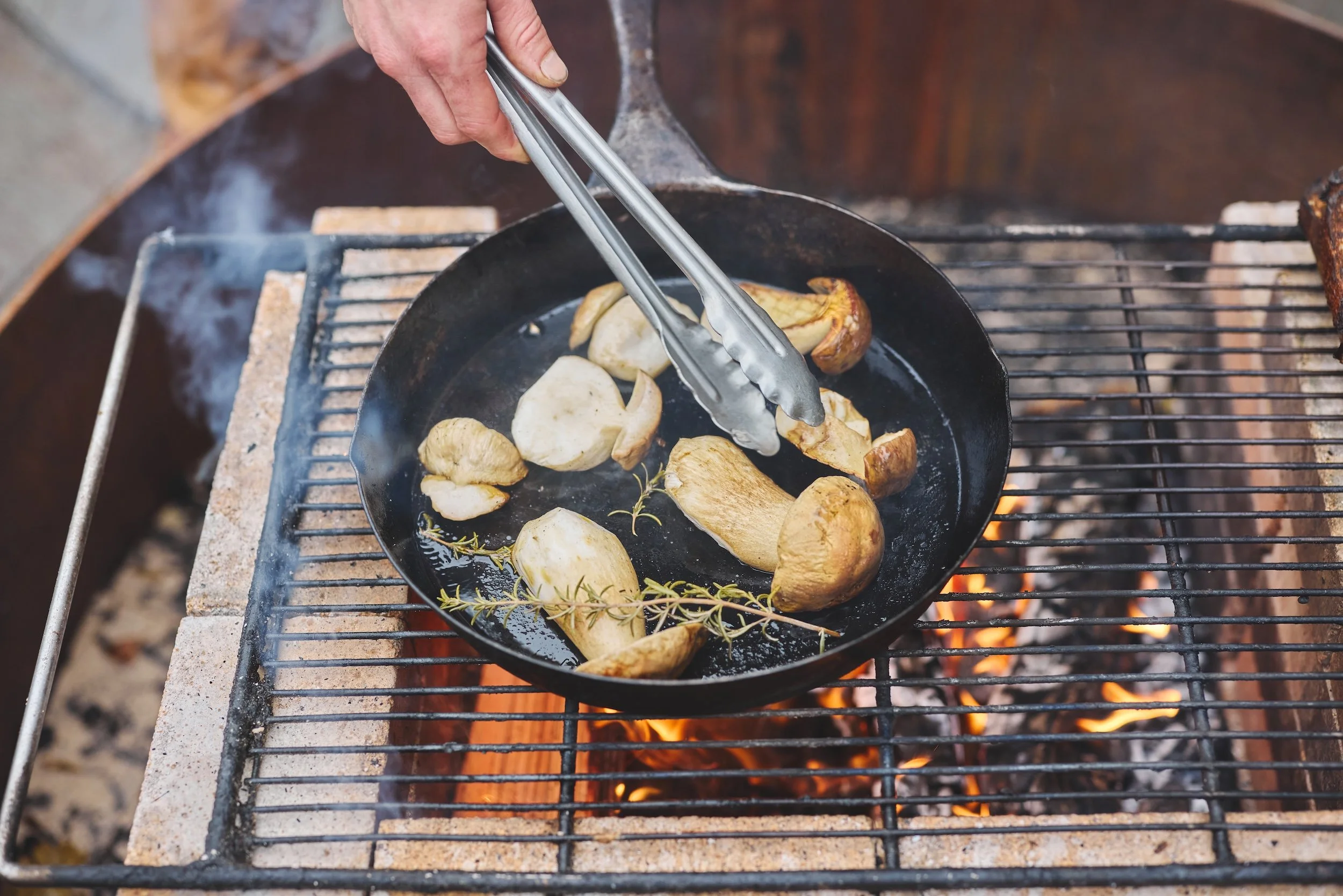 Fried mushrooms cooking in a black cast iron skillet over an open flame on a grill.