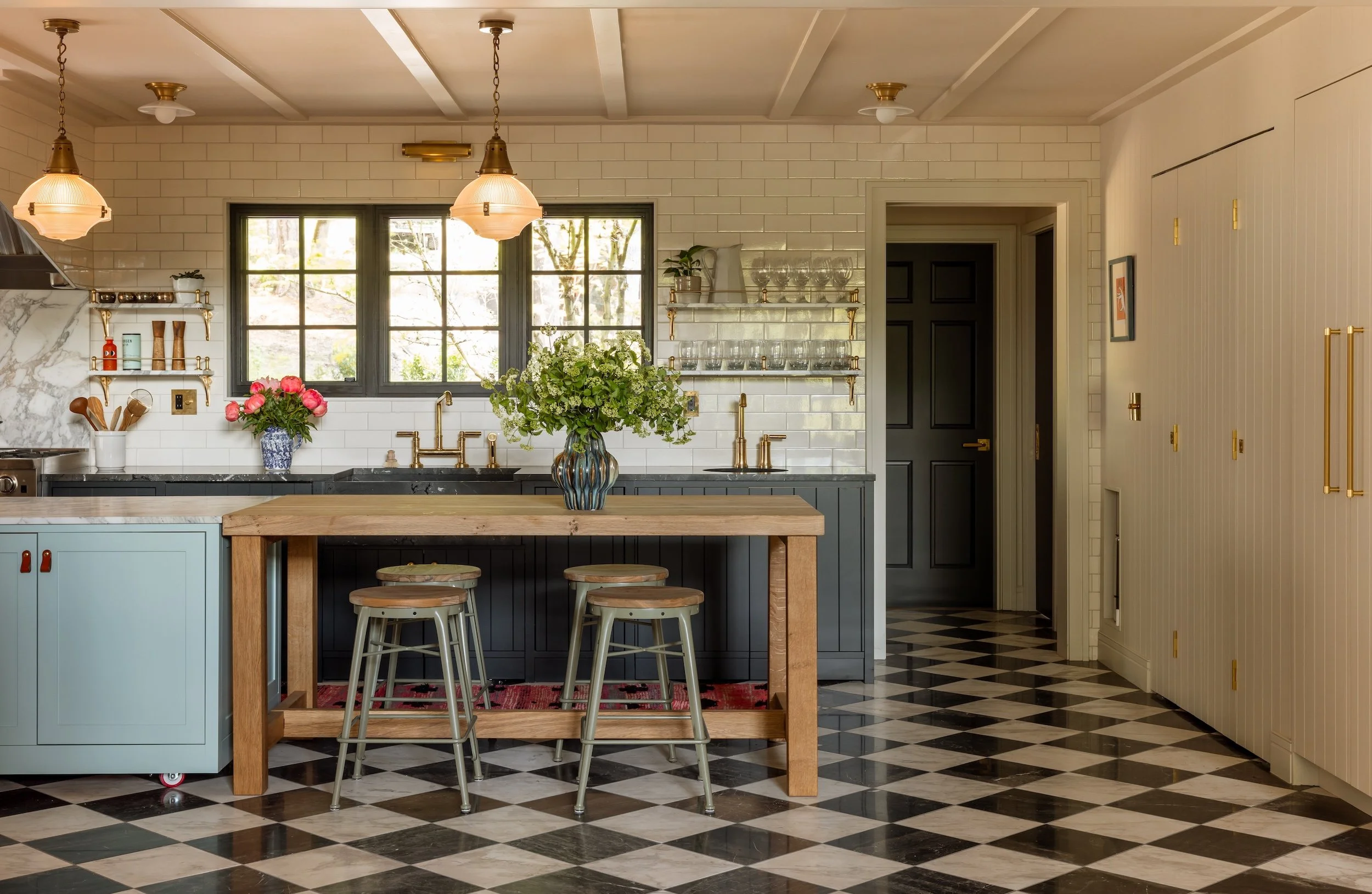 Kitchen with black and white checkered floor tiles, blue island with wooden countertop, two metal stools, blue cabinets, white tiled backsplash, window with trees outside, open shelves with glassware and decorative items, flowers in a blue and white 