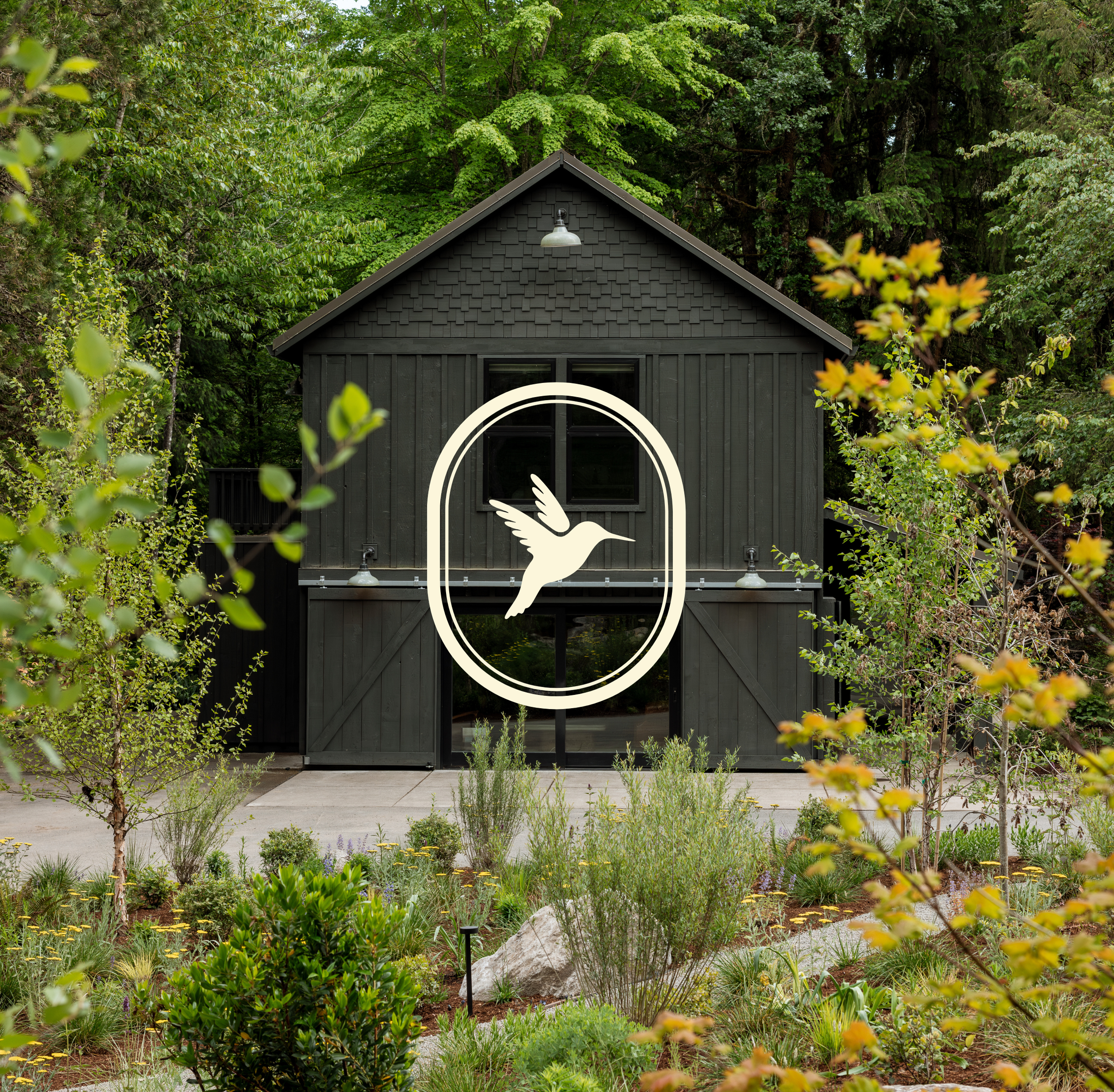 Black shed with a white bird emblem on the door, surrounded by lush green trees and a garden with various plants and flowers.