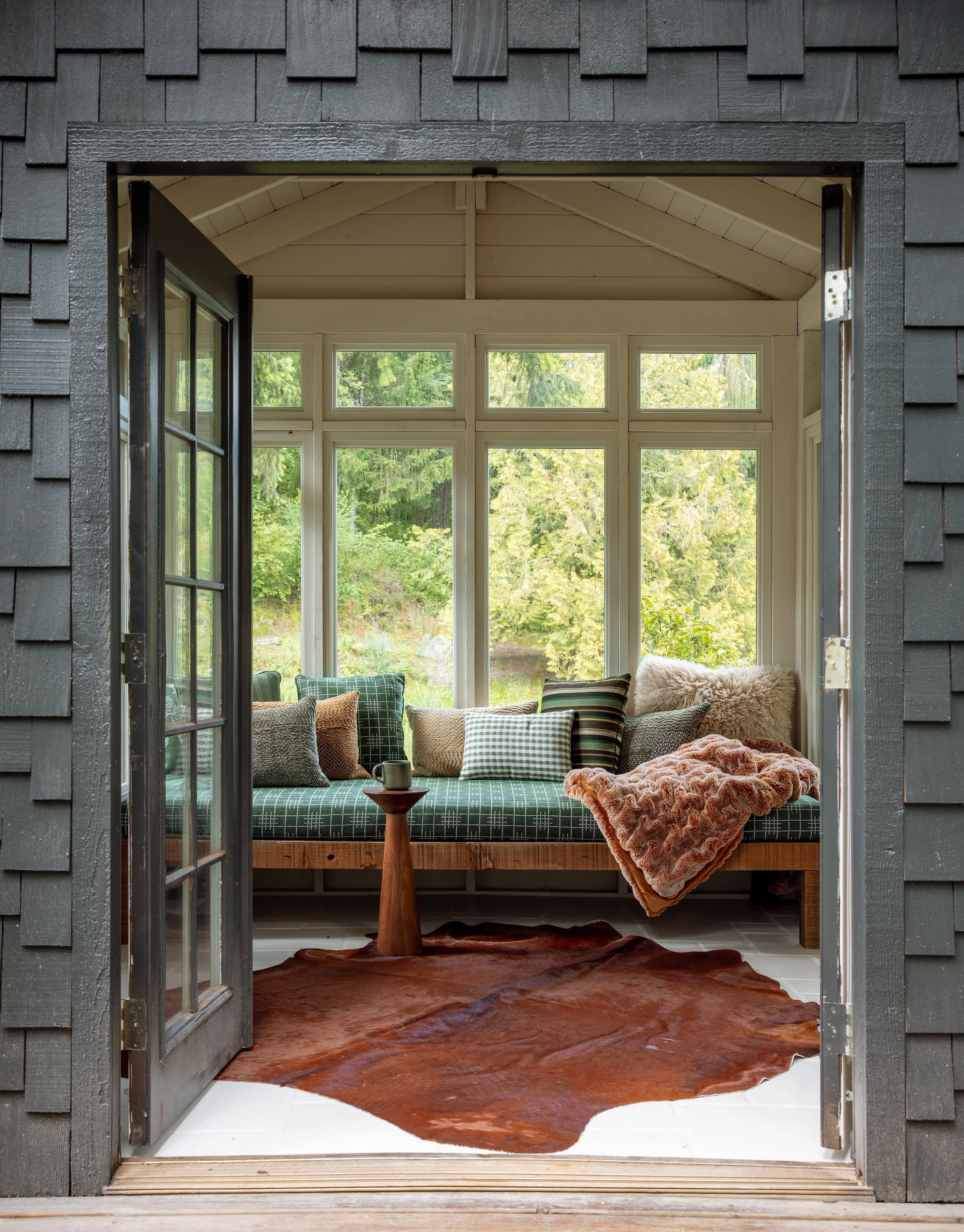 View through a dark gray shingled exterior wall opening into a cozy sunroom with large windows and a view of green trees outside. Inside, there is a sofa with an assortment of pillows and a blanket, a small wooden side table with a cup on it, and a l