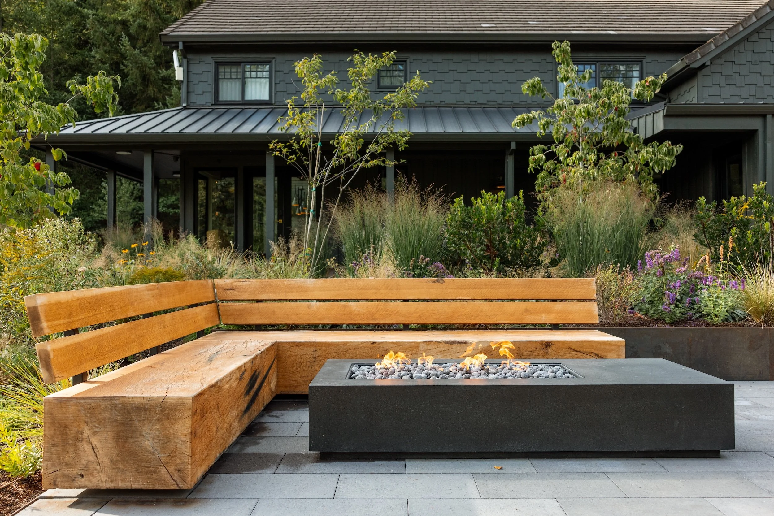 A backyard patio with a wooden bench, a fire pit with flames, and lush plants surrounding a dark-colored house with multiple windows and a metal roof.