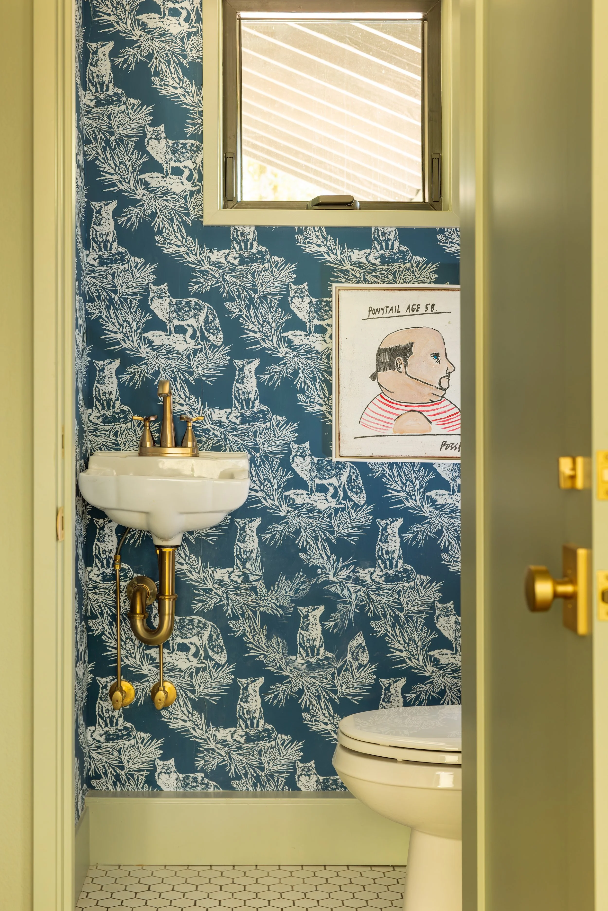 A small bathroom with wallpaper featuring white wolves and trees on a blue background. There is a white sink with gold fixtures, a mirror, and a framed drawing of a man with the caption 'Ponytail Age 58' above a toilet with a closed lid. A small window is above the mirror.